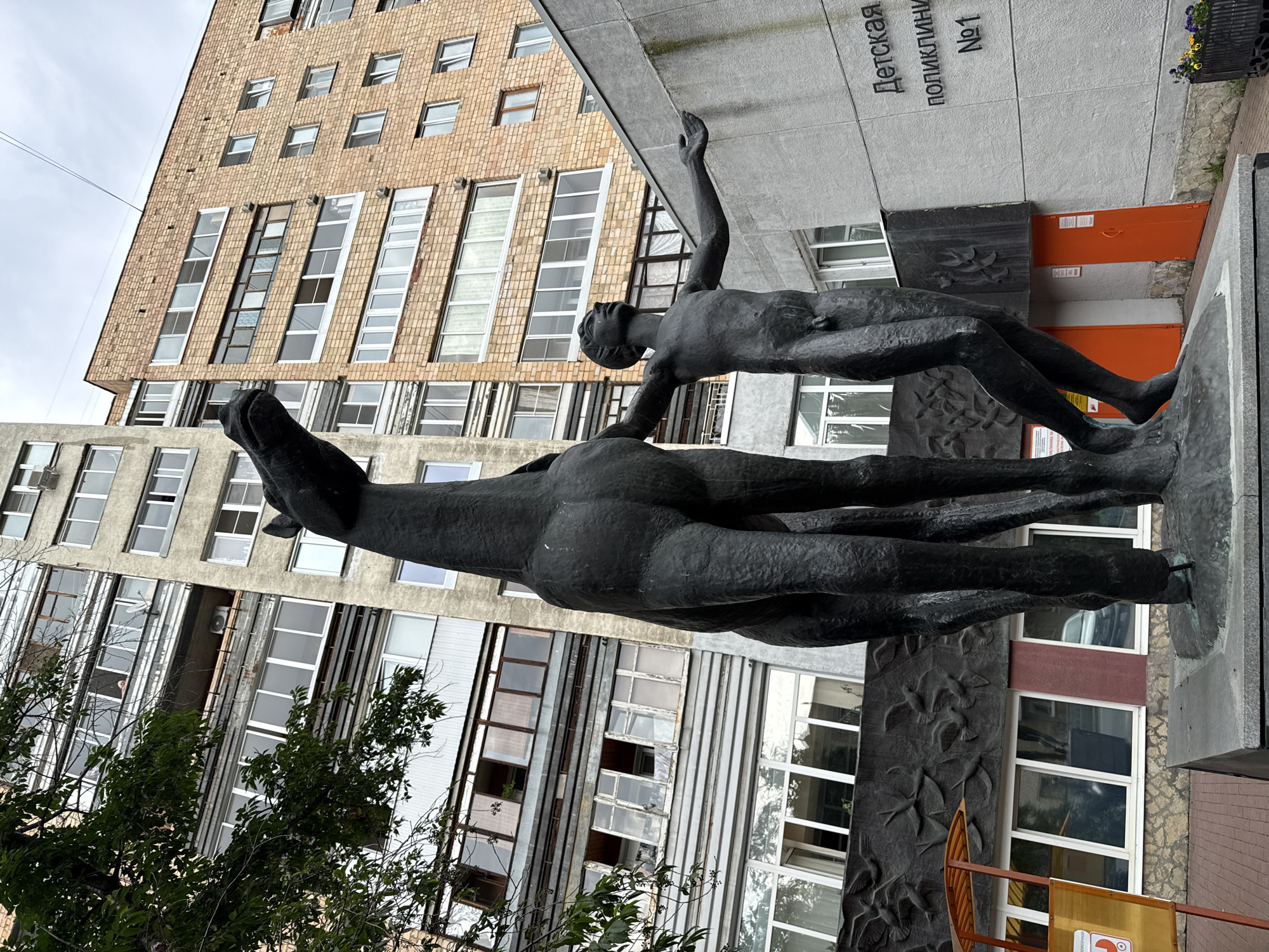 The image shows a statue of a boy reaching towards a horse in front of a brick building with multiple windows and balconies. The statue is made of dark metal and stands on a pedestal. The building has a sign that reads "Детская поликлиника №1" in Russian, which translates to "Children's Polyclinic No. 1". The statue is located in Yekaterinburg, Russia, specifically at Children's Polyclinic No. 1 on Krasnoarmeyskaya Street. The weather appears overcast.