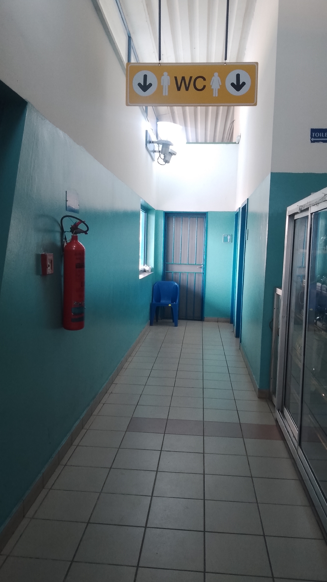 A hallway in Orerokpe, Nigeria, features a red fire extinguisher mounted on a turquoise wall on the left. The floor is tiled in a light-colored grid pattern. A blue plastic chair sits against the wall near a window and a metal door at the end of the hallway. A yellow sign hangs from the ceiling indicating the direction of restrooms (WC). A cooler with glass doors is on the right side of the hallway.
