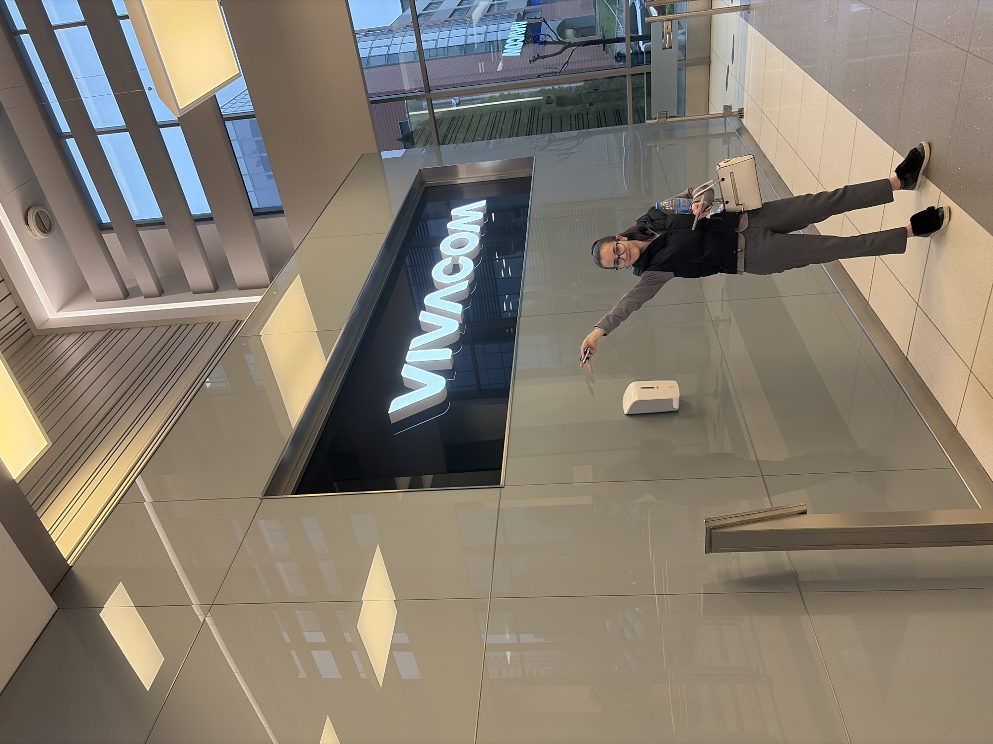 A person wearing glasses, a gray shirt, and black shoes is standing at an angle on a tile floor with a white handbag to their left, pointing with their right hand toward a white wall-mounted object. To the left of the person, a large sign with the word "VIVACOM" is illuminated. The building's interior includes a ceiling with multiple light fixtures and windows showing another building across the street. Based on the "VIVACOM" signage, the location is likely in Sofia, Bulgaria.
