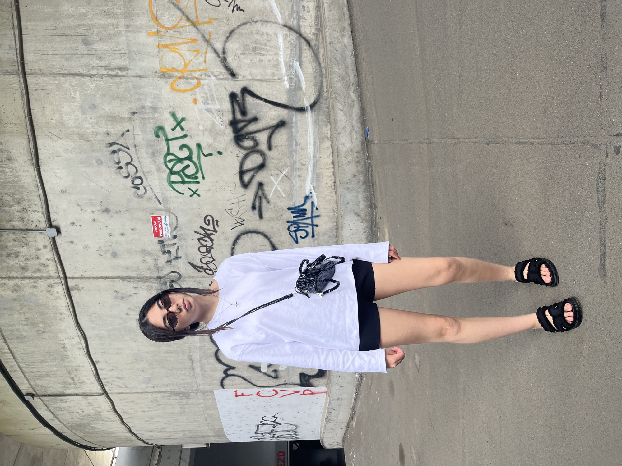 A young woman stands in front of a concrete wall covered in graffiti. She is wearing a white long-sleeved shirt, black shorts, black sandals, and sunglasses. A small black crossbody bag is slung over her shoulder. The concrete wall has various graffiti tags in different colors, including red, green, yellow, and black. To the left of the woman, a section of a building with an entrance is visible. The ground is paved with asphalt. The location is identified as Pilsen, Czechia.