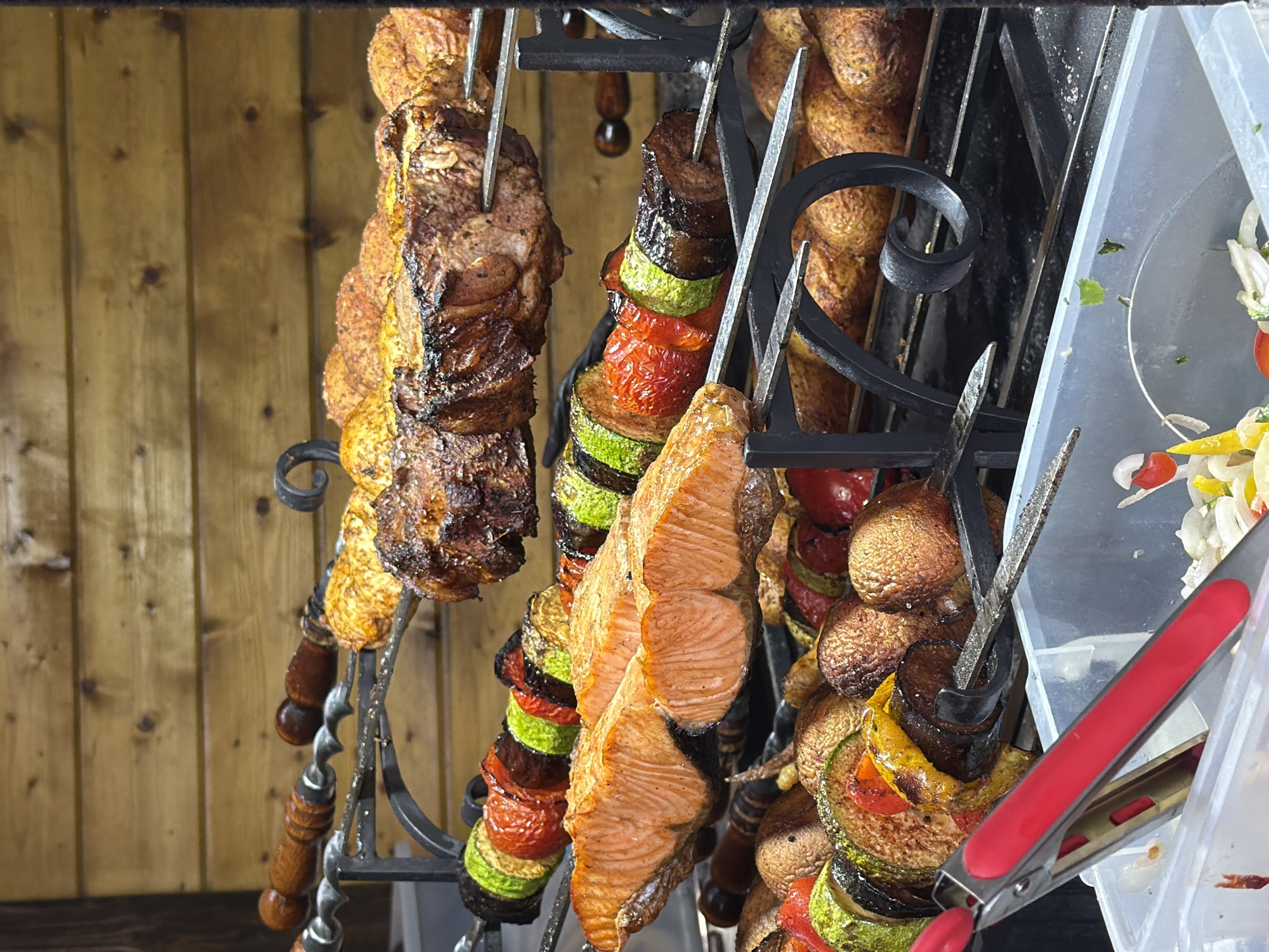 The image shows several skewers of grilled food, including salmon, meat, and vegetables, being cooked over a grill. The food is arranged on metal skewers and is displayed in front of a wooden background. In the foreground, there is a clear plastic container holding some salad ingredients, and a pair of red and silver tongs. The setting appears to be an outdoor food stall or market. This photograph was taken in Sochi, Russia.