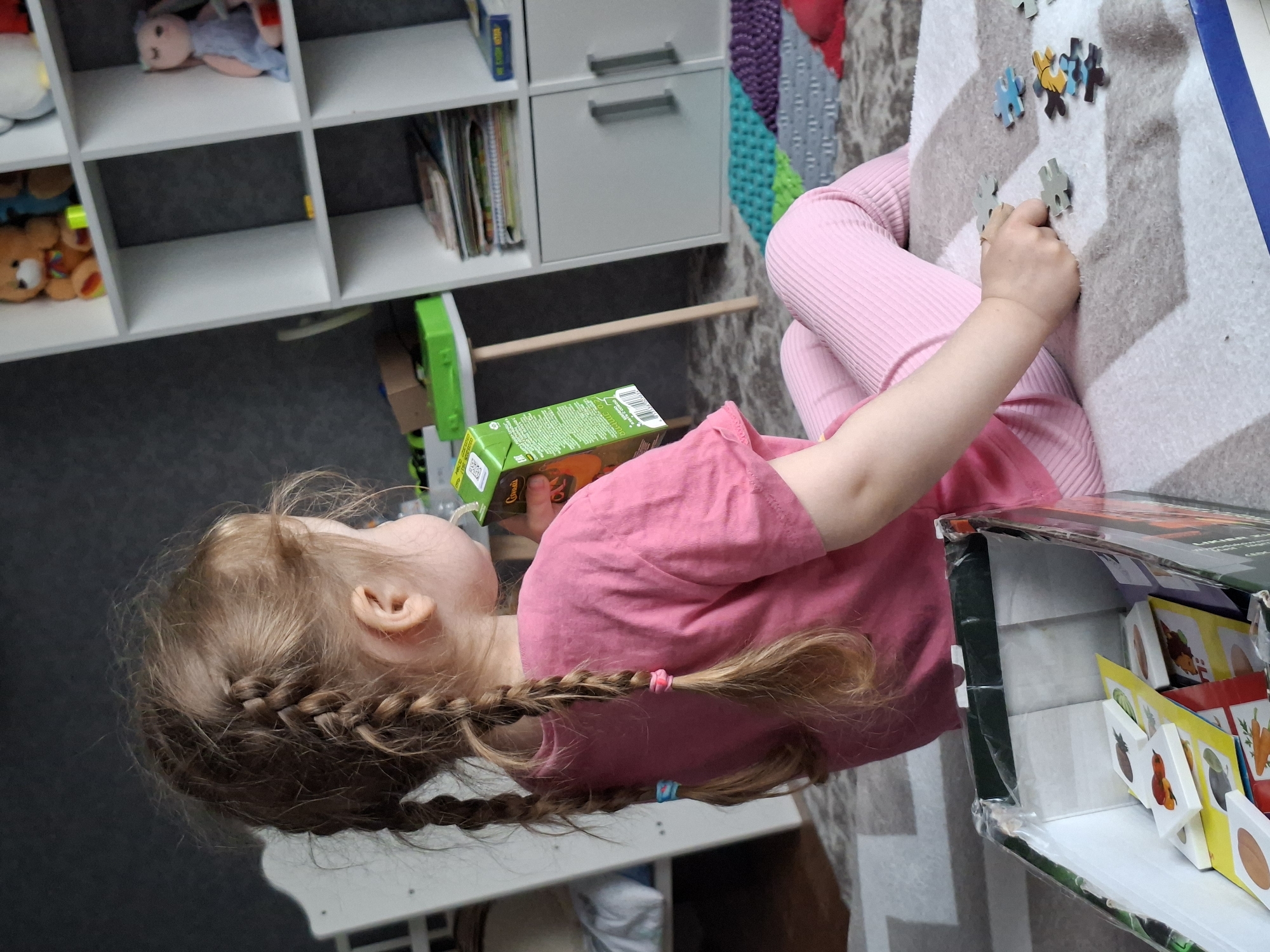 The photograph captures a young girl with braided hair, wearing a pink shirt and leggings, sitting on a patterned rug. She is engaged in putting together a jigsaw puzzle, with puzzle pieces scattered on the rug in front of her. She is also drinking juice from a green juice box. The setting appears to be indoors, possibly in a playroom or living area, with a white shelving unit filled with toys and books in the background.
