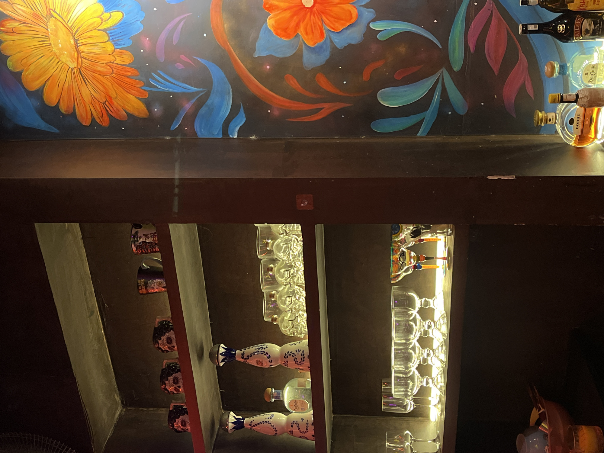 The image depicts a bar in Đà Nẵng, Vietnam. The upper portion shows a colorful mural with floral designs. On a shelf above the bar, there are various bottles of alcohol. Below the bar counter, shelves display rows of wine glasses, skull-shaped bottles, and intricately decorated bottles, possibly containing tequila.