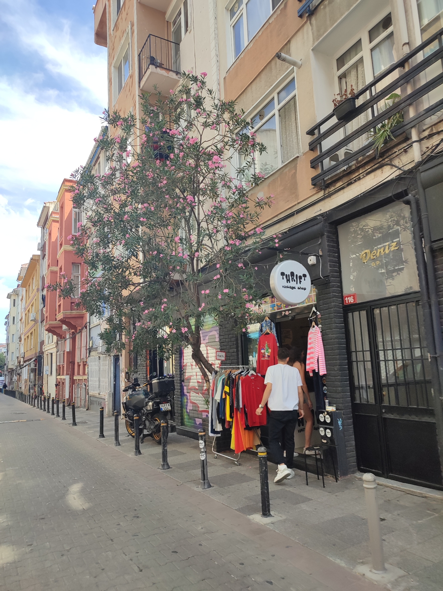 The image depicts a street scene in Kadıköy, Turkey, on a cloudy day. In the foreground, a cobblestone sidewalk lines the street, with black bollards marking the edge. A motorcycle is parked on the right side of the sidewalk. Further down the street, a vintage clothing shop named "Thrift" is visible, with clothing racks displayed outside. A young man in a white t-shirt and black pants, and a young woman in a black outfit are walking past the shop. Behind the shop, a building with multiple stories and balconies can be seen. The street is lined with colorful buildings, giving it a vibrant, urban feel.
