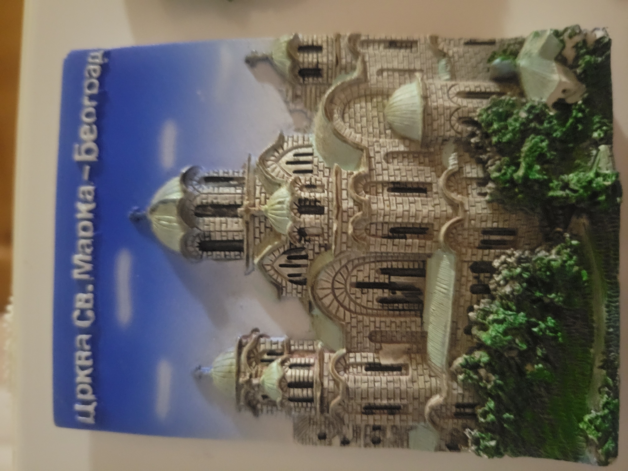 This is a close-up, eye-level, medium shot of a souvenir magnet depicting a church. The text on the magnet reads "Црква Св. Марка - Београд", which translates to "Church of St. Mark - Belgrade". The church is made of light-colored bricks with green domes. It is a detailed miniature with multiple towers and arched windows. In the foreground, there are green trees and grass, representing a landscape. The background is a blue sky with white clouds. The magnet is placed on a light-colored, textured surface. The overall impression is that of a tourist souvenir. The mention of Belgrade, Serbia, indicates the location of the depicted church, not Belaazyorsk, Belarus.
