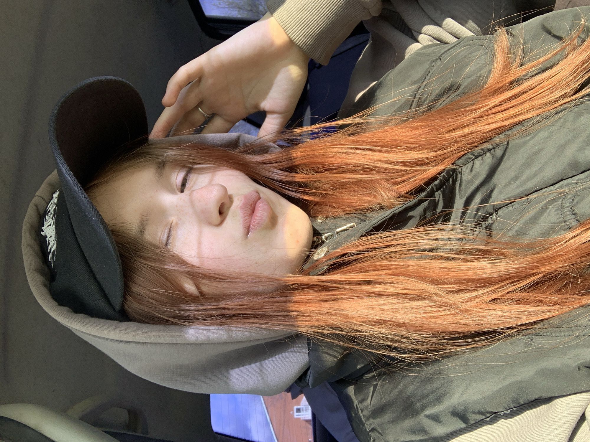 A young person with long, vibrant reddish-auburn hair is captured in a relaxed pose, possibly lying down. They are wearing a black baseball cap, a light beige/grey hoodie, and a dark green/grey puffer jacket. Subtle freckles are visible on their face, and their eyes are either partially closed or looking downward. Their lips are slightly parted and appear soft. One hand, adorned with a simple silver ring on the ring finger, is gently raised, resting near their head and the brim of their cap.
Bright, natural sunlight streams from the right side of the frame, casting strong highlights on their hair and face, and creating distinct shadows, indicating it is daytime. The background is blurred but suggests an indoor setting, possibly the interior of a vehicle due to the visible textures. The overall impression is one of calm, rest, or peaceful contemplation. No visible text is present in the image. The location context is Фоки, Russia.