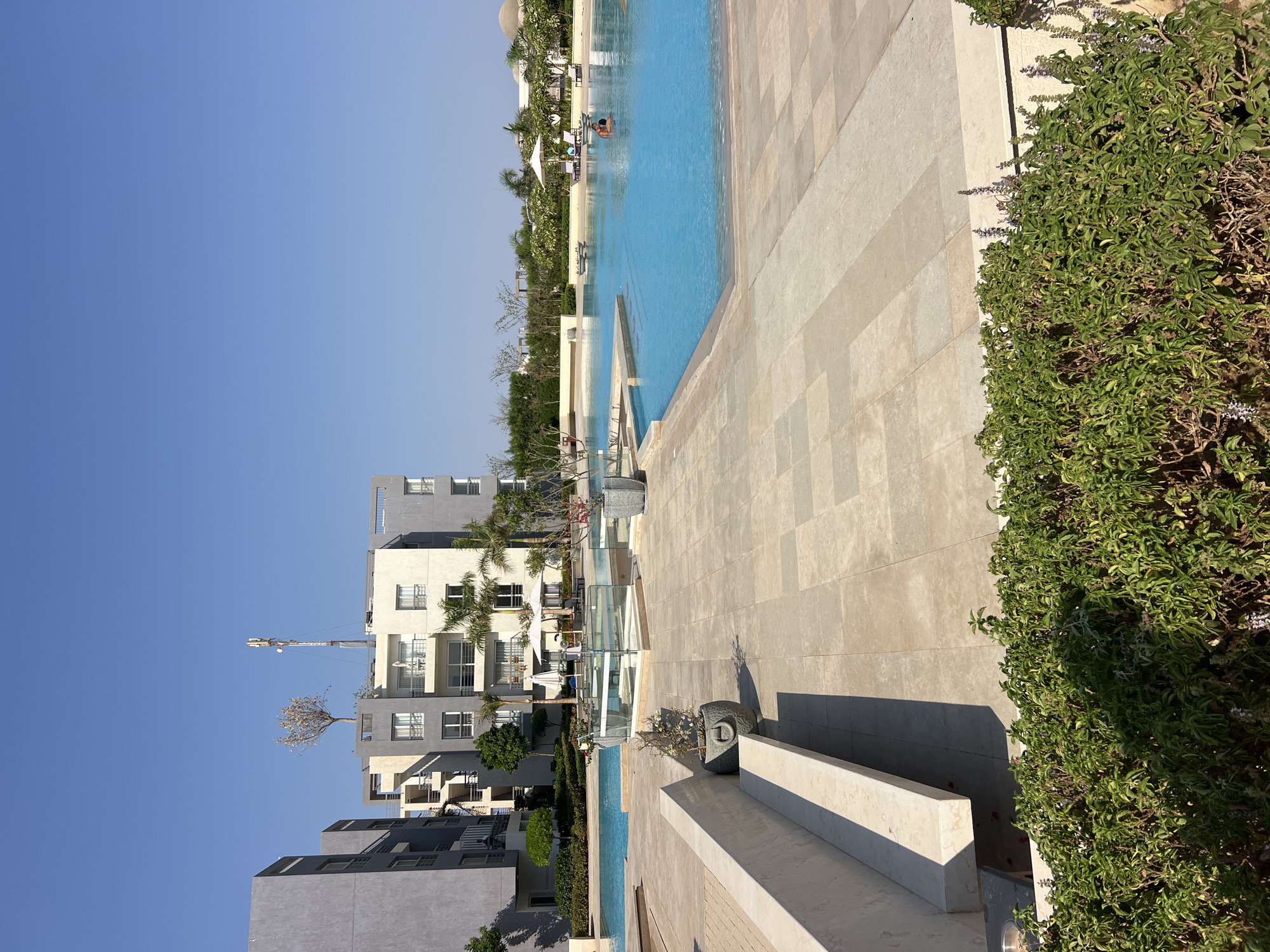 The image depicts an outdoor scene at a resort in an unknown city in Egypt. In the foreground, a paved area with large, light-colored square tiles leads to a long, multi-tiered concrete bench. Lush green bushes with some flowering plants are positioned in front of the bench. Beyond the paved area, a large, bright blue swimming pool occupies the midground and extends to the right. Several lounge chairs and umbrellas are visible around the pool. In the water, two individuals are present. In the background, there are modern, multi-story buildings with a mix of gray, white, and beige facades. One building has a tree growing on its roof and a prominent communication tower. Palm trees and other greenery are scattered throughout the resort. The sky is clear and a vibrant blue, indicating a sunny day. The overall atmosphere suggests a leisure and relaxation environment.