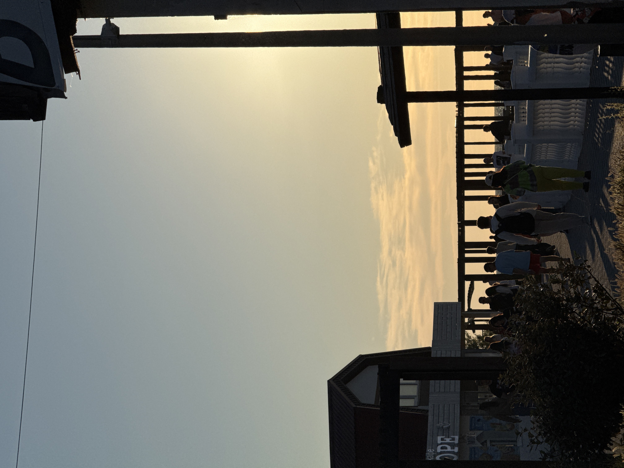 The image depicts a bustling scene at dusk, likely a boardwalk or promenade, with a clear sky transitioning from a pale blue to a warm yellow hue as the sun sets. A silhouetted structure, possibly a signpost with the letter "D" visible, stands prominently on the right, casting a long shadow. To the left, a building with a dark roof and white siding is partially visible, with a sign that appears to read "HOPE".
A diverse group of people are strolling along a paved pathway lined with ornate white railings. The individuals are captured in various poses, some walking forward, others conversing, and some are silhouetted against the fading light. The atmosphere suggests a leisurely evening activity, with the sea or a large body of water visible in the distance behind a wooden pergola. The foreground is partially obscured by dark foliage, adding depth to the scene. The overall impression is one of a lively public space enjoying the end of a day.