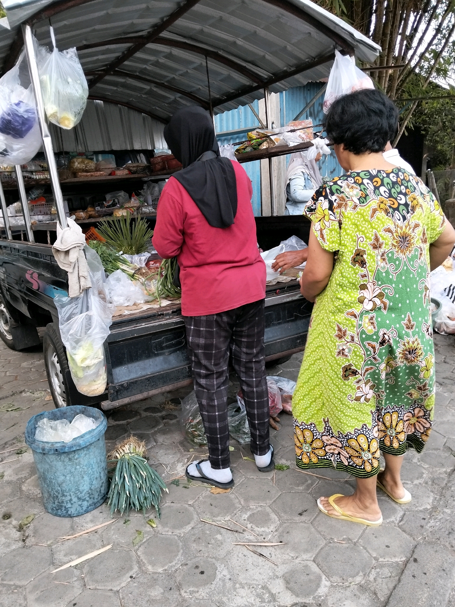 Two women are standing at the back of a pickup truck that is parked on a paved street in Pangenjurutengah, Indonesia. The truck has a metal canopy and shelves filled with goods. The woman on the left is wearing a red shirt, black headscarf, and plaid pants. She is holding a bunch of green onions. The woman on the right is wearing a green dress with a floral pattern. She is looking at the woman on the left and holding a plastic bag. There is a blue bucket and more green onions on the ground next to the truck. The area appears to be a market or street vendor stall.