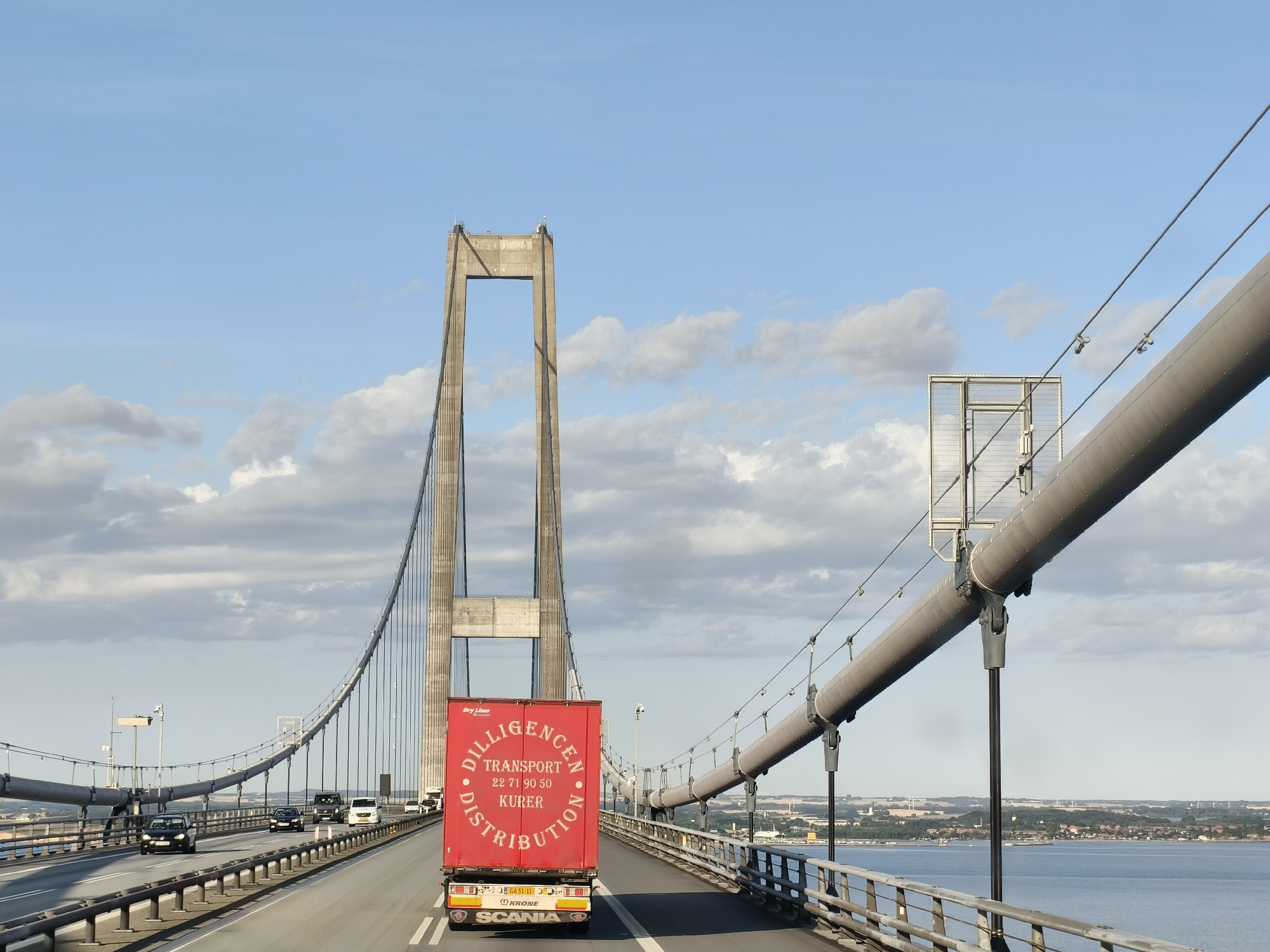 The image shows a view from a car on a suspension bridge in Unknown, Denmark. The bridge has two large concrete towers, with cables suspending the roadway. A red semi-trailer truck with "DILLIGENCEN TRANSPORT" written on it is in the foreground, driving away from the camera. Several other cars are also on the bridge. The sky is blue with scattered clouds, and the sea is visible on the right side of the frame. In the distance, a cityscape with buildings and windmills can be seen on the horizon. The overall scene depicts a typical day of travel on a major bridge.