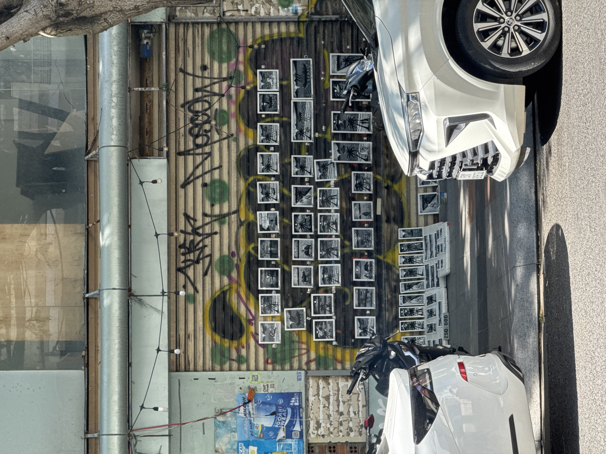 The image depicts a street scene in Nha Trang, Vietnam, focusing on a storefront converted into an outdoor art display. The central element is a closed, corrugated metal roller shutter, heavily spray-painted with black, yellow, and green graffiti, prominently featuring the word "MOSCOW." Approximately 30 small, framed, black-and-white art prints, depicting various tropical and rural Vietnamese scenes such as palm trees, buffalo, boats, and figures, are affixed directly to this shutter. Below, on the concrete sidewalk, an additional 20 or more similar unframed prints are displayed, accompanied by a price sign indicating "250.000."
To the left of the shutter, a faded light blue painted wall section displays a partial advertisement for Pocari Sweat and exposed brickwork. Above the shutter, a horizontal metallic duct runs across the building facade, with a string of bare light bulbs suspended below it. Reflective windows and exposed structural elements are visible on the upper building. A large tree trunk is present on the far right, partially obscuring the upper building section.
Vehicles dominate the foreground. A white SUV, resembling a Toyota Avanza, is parked on the street in the right foreground, facing right, with its front grille, headlight, and passenger-side wheel visible. A black Honda scooter is parked directly in front of the art display, positioned between the white SUV and the rear section of another white car, partially visible on the far left. The scene is illuminated by strong natural daylight, casting distinct shadows on the asphalt street and concrete sidewalk. No individuals are present within the visible frame.