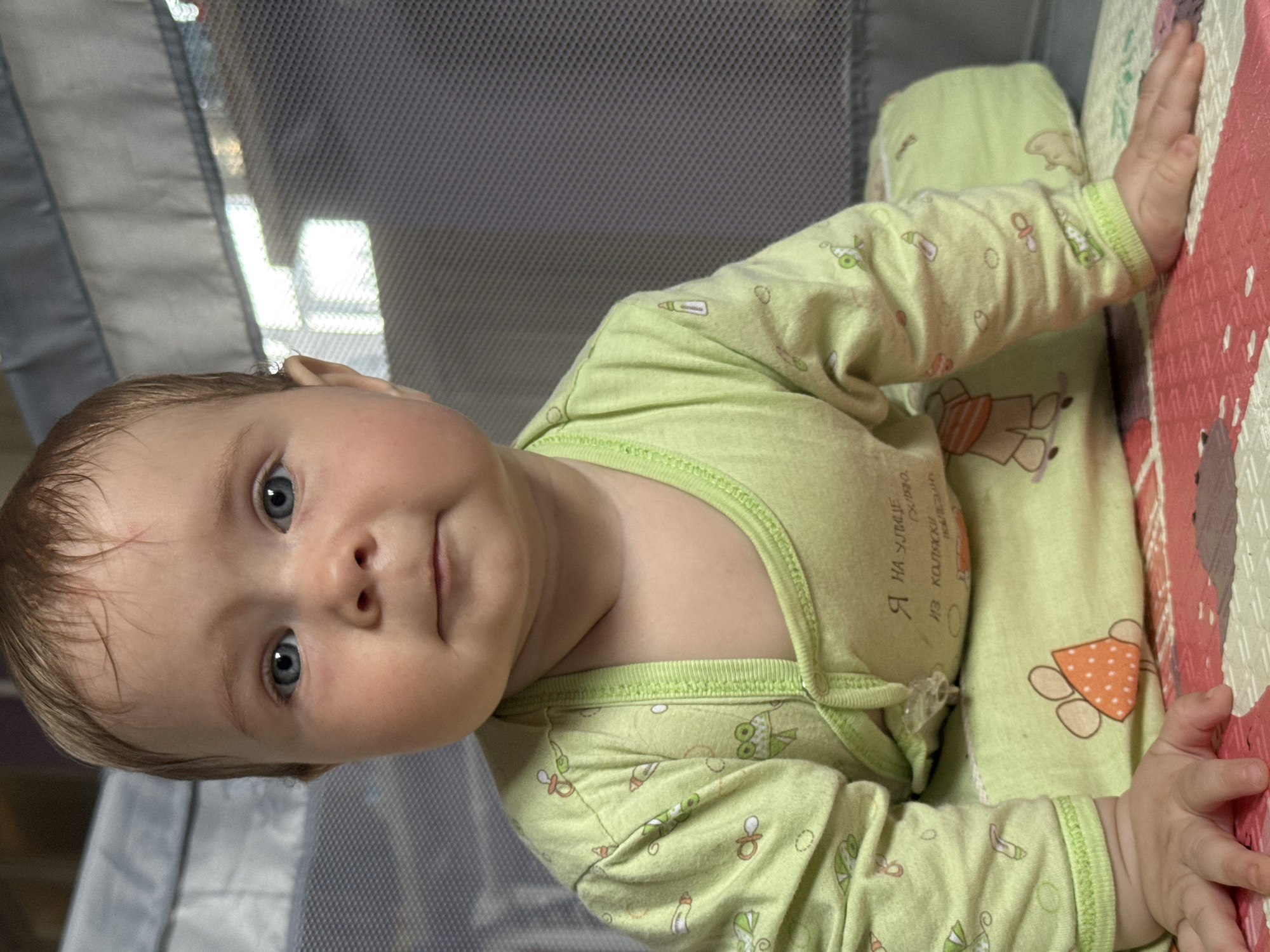 A baby with light brown hair and striking blue eyes lies in a playpen or portable crib, looking directly at the camera with a neutral to slightly curious expression. The baby is wearing a light green, long-sleeved bodysuit or sleepsuit, patterned with various light-colored illustrations including baby bottles, pacifiers, owls, and other cartoon figures. On the baby's torso, Cyrillic text is visible on the clothing, including "Я НА УЛИЦЕ" (I AM ON THE STREET) and "ИЗ КОЛЯСКИ" (FROM THE STROLLER).
The baby's hands are visible, with the right hand resting on a colorful playmat section featuring a red and white geometric or mosaic pattern. A lighter section of the mat under the baby's body also shows a cartoon animal figure, possibly a bear, with an orange belly. The background reveals the light grey mesh side of the enclosure, with bright reflections suggesting an indoor setting illuminated by natural light, likely from a window. The activity captured is the baby resting or engaging in gentle observation. The scene is set indoors in Rusalevka, Russia.