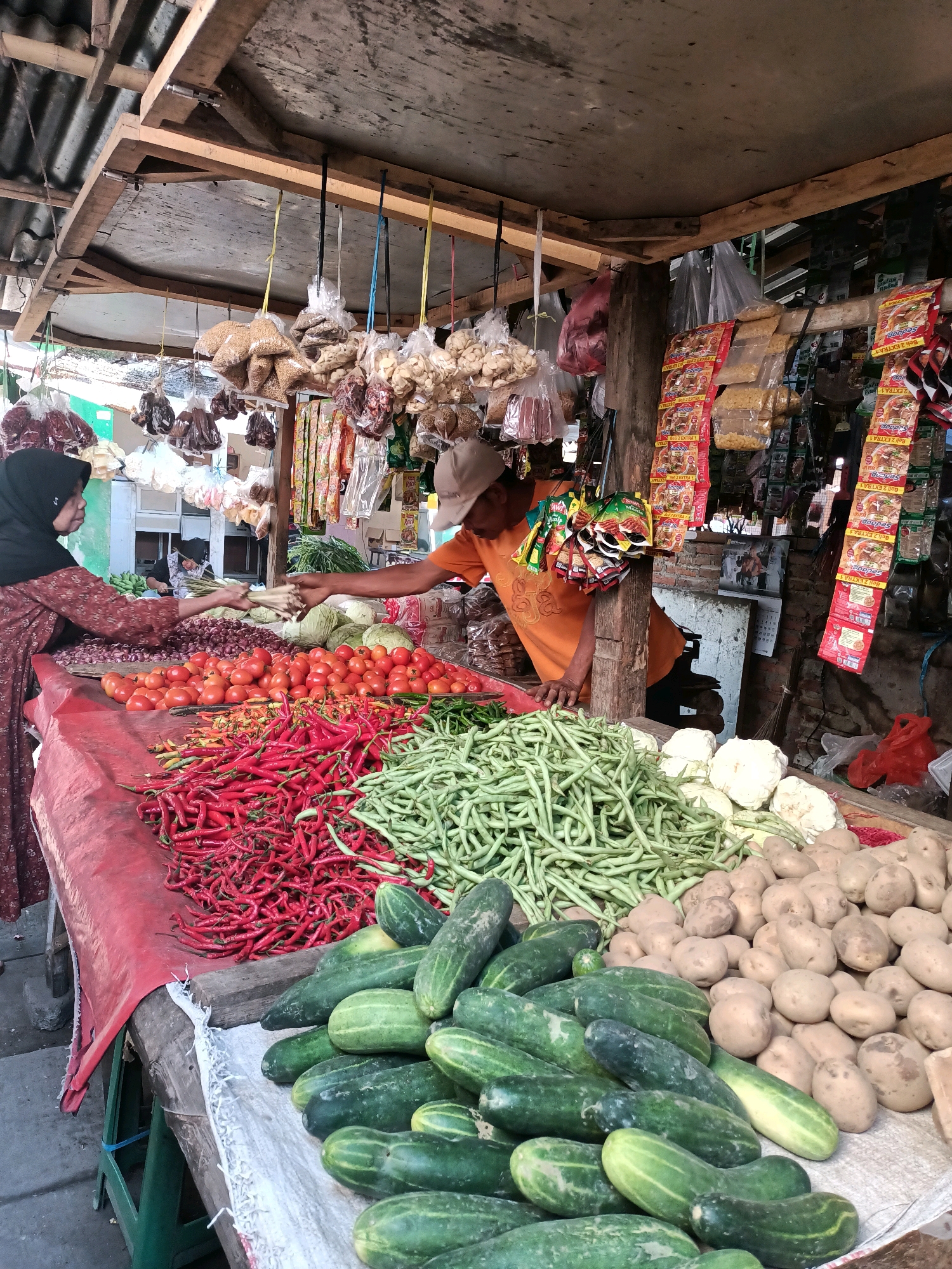 A street vendor in Badas, Indonesia, arranges a stall piled high with fresh produce. A woman wearing a hijab and a patterned dress selects vegetables from the stall, extending her hand to a vendor in an orange shirt and a cap. The stall is covered by a wooden awning, with goods hanging from the ceiling and stacked on tables. In the foreground, a variety of vegetables are displayed, including red chilies, tomatoes, green beans, cucumbers, potatoes, and cauliflower. Bags of dried goods and packaged food items hang from the stall's structure. The ground is made of rough pavement.
