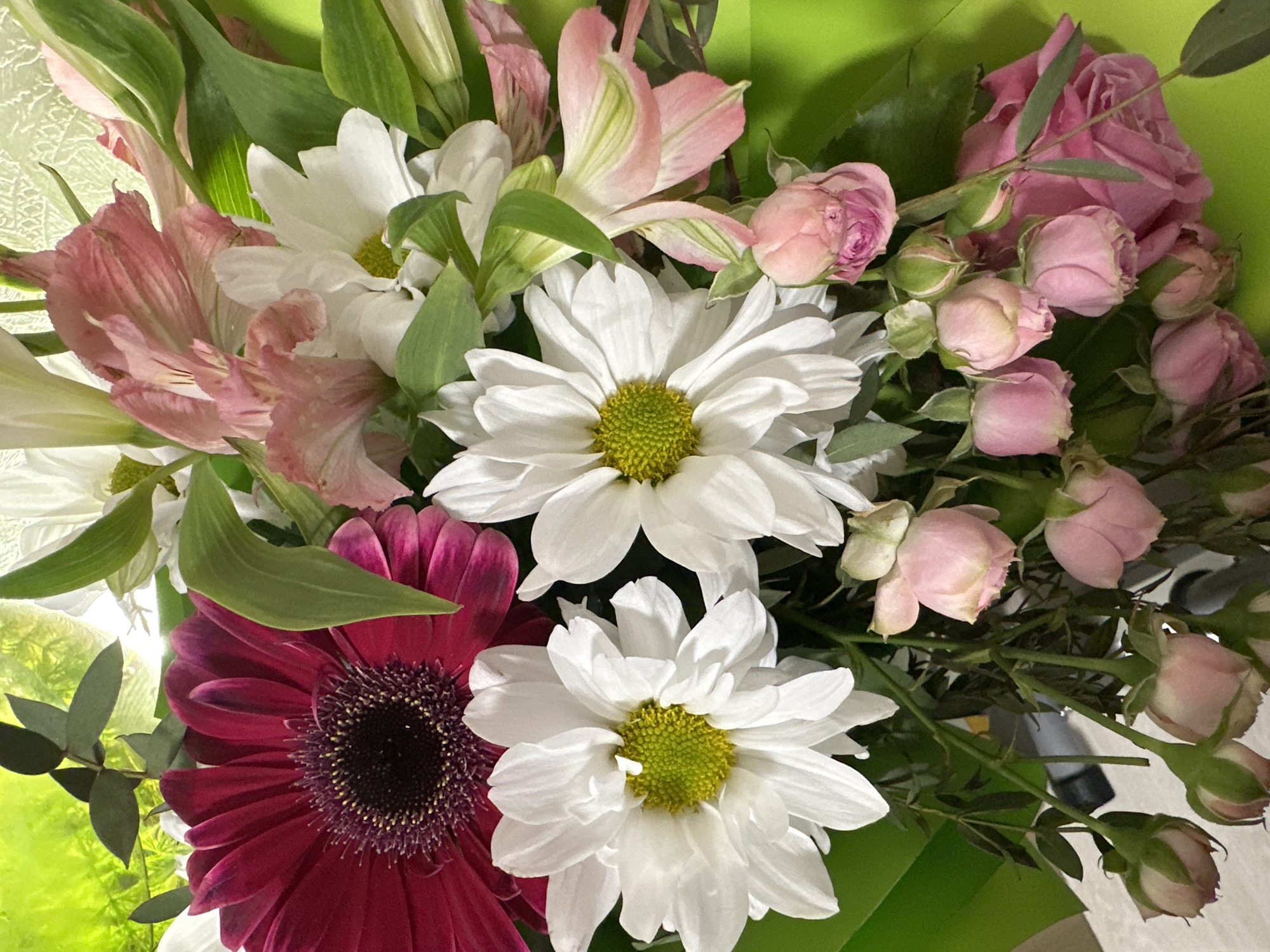 The image shows a close-up of a bouquet of flowers. The bouquet includes pink alstroemeria, white daisies with yellow centers, pink roses, and a burgundy gerbera daisy with a dark center. The flowers are arranged with green leaves and stems visible. The background is a light green color. There is no information in the image to identify the location as the city of Uva, Russia.