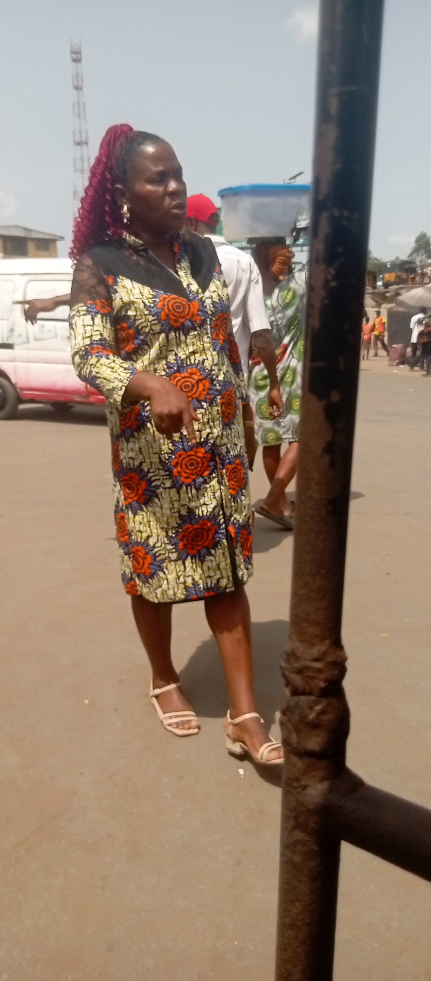 The image depicts a woman standing on a street in Onitsha, Nigeria. She is wearing a knee-length dress with a floral pattern and black lace sleeves. Her hair is styled in a high ponytail with burgundy extensions. She is wearing sandals and pointing her finger downward. In the background, there are other people, a white van, and a metal pole. The street appears to be a busy thoroughfare.