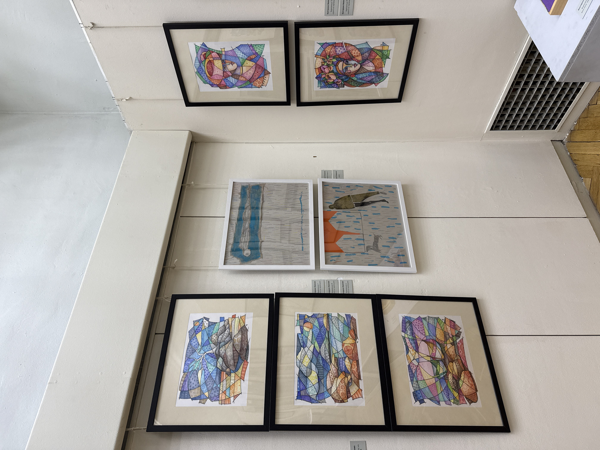 The image shows a vertical arrangement of framed artwork displayed on a white wall within a building in Pazardzhik, Bulgaria. The wall has visible wiring running horizontally across it to support the frames. On the top row, two framed drawings depict stylized female faces with colorful, patterned hair. The middle row features two framed pieces with white frames: one depicting blue vertical lines on a gray background, and another depicting an orange and gray figure swimming in a blue striped area. The bottom row contains three framed drawings with black frames, each featuring an abstract design with colorful geometric shapes. Small labels or plaques are visible below some of the frames. An air vent is present near the top right corner. The wall appears to be part of an exhibition or display area.