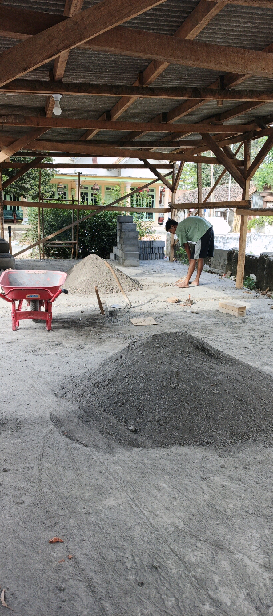A low-angle, vertical, full shot captures a construction scene in Kasiyan, Indonesia, under a wooden-framed, corrugated metal roof structure. A young man wearing a green t-shirt and black shorts is the sole human subject, bent over and sweeping a small pile of dirt or gravel on the concrete ground with a broom. The environment is utilitarian and shows signs of ongoing construction. In the foreground, a large mound of gray gravel and sand dominates the lower portion of the frame. To the left, a red wheelbarrow is parked, facing the viewer, with a small pile of the same gray material beside it. Two garden spades are propped upright near a smaller pile of sand further back. In the middle ground, a stack of gray concrete blocks stands to the right of the central sand pile. Beyond the construction area, a traditional Indonesian building with light green trim and windows is visible, surrounded by lush green trees. The ground is a mix of compacted dirt and concrete, with tire tracks imprinted on the surface, indicating recent vehicle activity. The overall impression is one of a functional, outdoor workspace in a rural or semi-rural Indonesian setting.