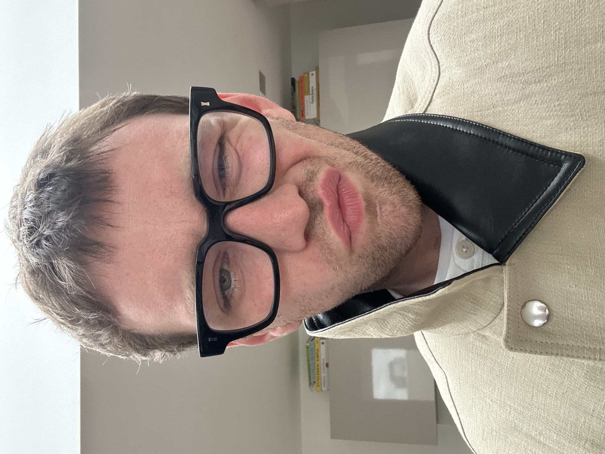 A close-up portrait shows a light-skinned male wearing black-framed glasses. He has short, light-colored hair and stubble. He is wearing a light-colored jacket with a black collar and a visible white button-down shirt beneath. The background appears to be a plain, light-colored wall. There is no evidence to suggest that the photo was taken in the city of London, United Kingdom.