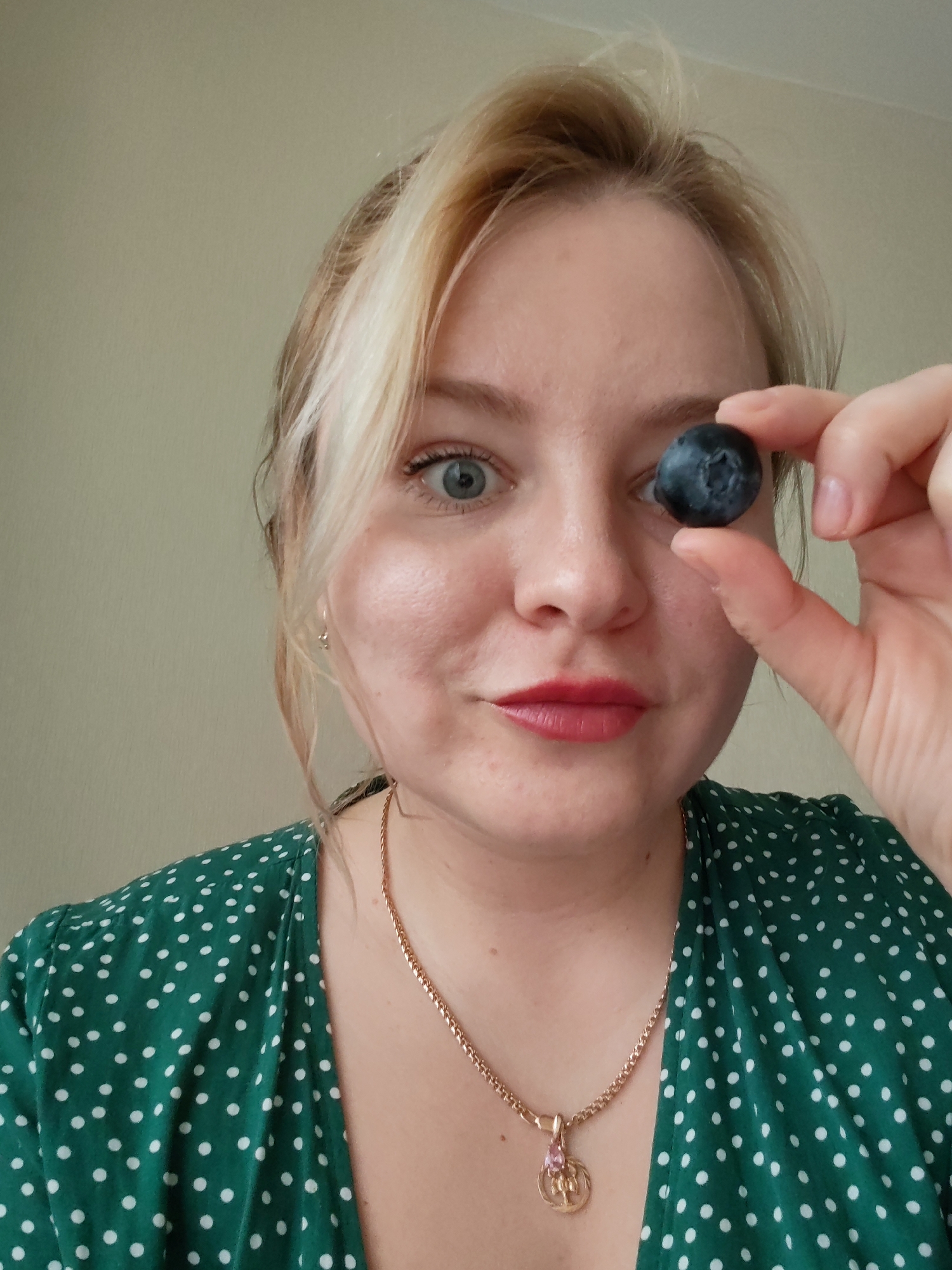 A young adult woman is captured in a playful, medium close-up shot indoors in Izhevsk, Russia. She has blonde hair with lighter highlights, styled loosely around her face. Her left eye, wide open and light blue or grey, looks directly at the viewer, while her right eye is obscured by a single, large blueberry that she holds between the thumb and forefinger of her right hand.
The woman has fair skin, with a light blush on her cheeks, and her lips are painted with red lipstick, hinting at a slight smile or smirk. She wears a green dress adorned with white polka dots and a gold chain necklace with a distinctive pendant featuring a pink stone and an intricate gold design. The background is a plain, light-colored wall, likely beige or pale green, providing a simple setting that keeps the focus on her and the blueberry. The lighting is soft and even, suggesting an indoor environment without harsh shadows. No text is visible in the image.