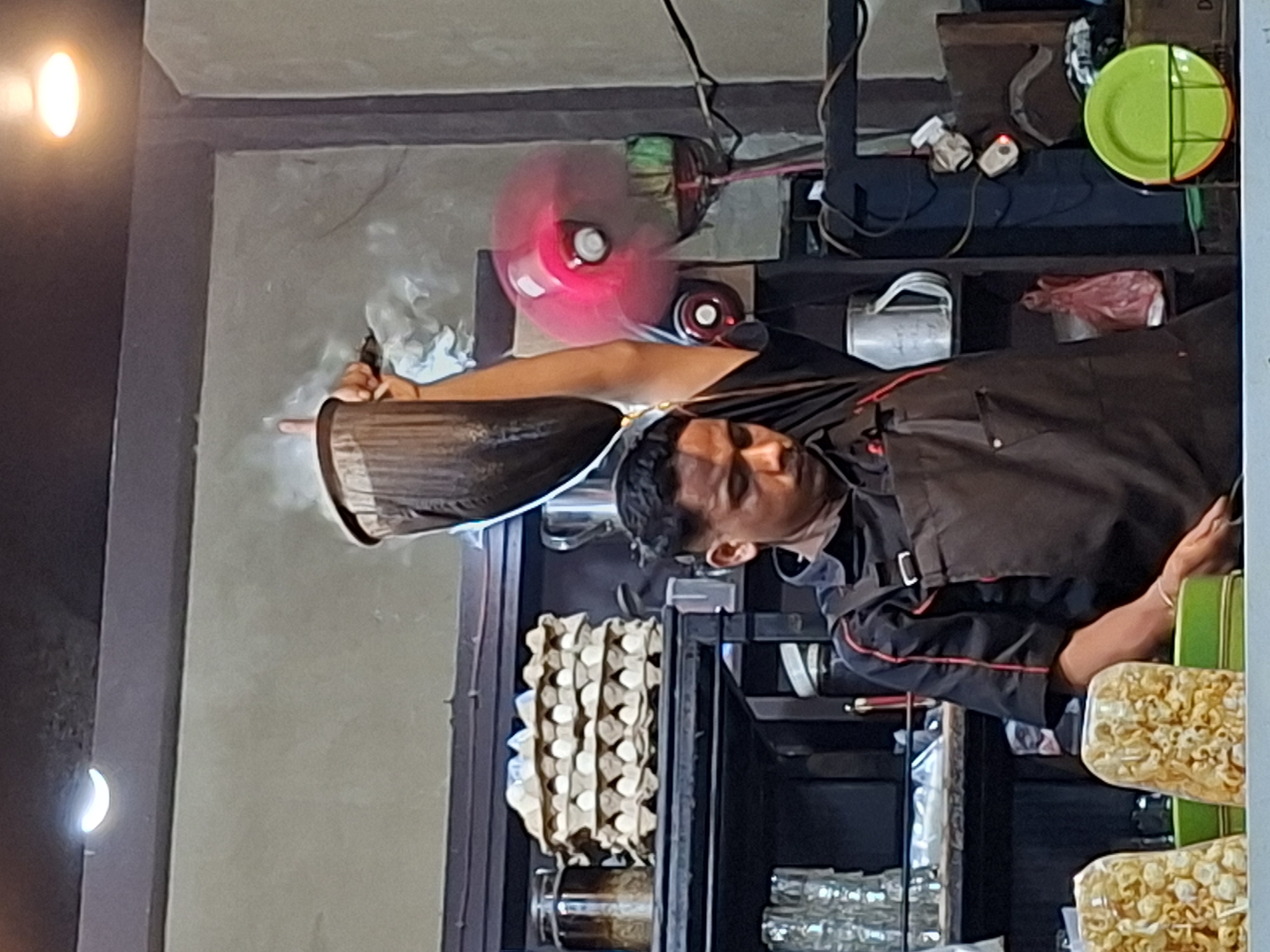 In an indoor setting, a man wearing a dark apron with red trim is positioned behind a counter. He is actively engaged in cooking or preparing food, as smoke emanates from the wok he holds. Behind him, there's shelving filled with kitchen supplies, an egg carton, glassware, and packaged food items. A small fan is visible in the background. This scene is presumably located in a kitchen or food preparation area in Lhokseumawe, Indonesia.