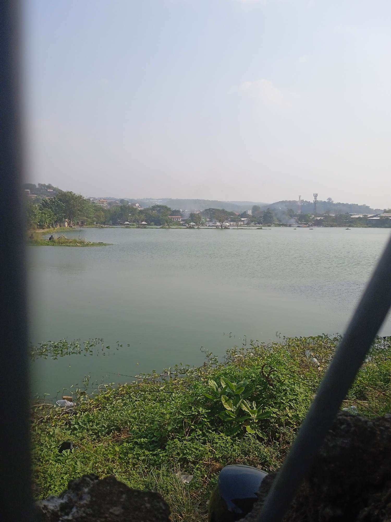 This is an eye-level, vertical, full shot of a serene lake on a hazy day, likely situated in Semarang, Indonesia. The foreground is dominated by lush green vegetation and some dark, rocky ground, partially obscured by a dark, vertical object on the left and a gray, diagonal pole on the right, suggesting a viewpoint from behind a fence or railing. The lake itself takes up a significant portion of the frame, its surface a muted green, reflecting the overcast sky.
In the mid-ground, a narrow strip of land lines the far side of the lake, covered with trees and some scattered buildings that extend up a gentle incline. Small white structures, possibly gazebos or huts, are visible near the water's edge, and a few small boats can be seen on the lake. On the left bank, a lone figure is seated on a small island of vegetation, fishing. The background features more distant buildings and hills, with a prominent cellular tower rising above the landscape. The overall atmosphere is tranquil, with the hazy sky softening the details of the distant city.