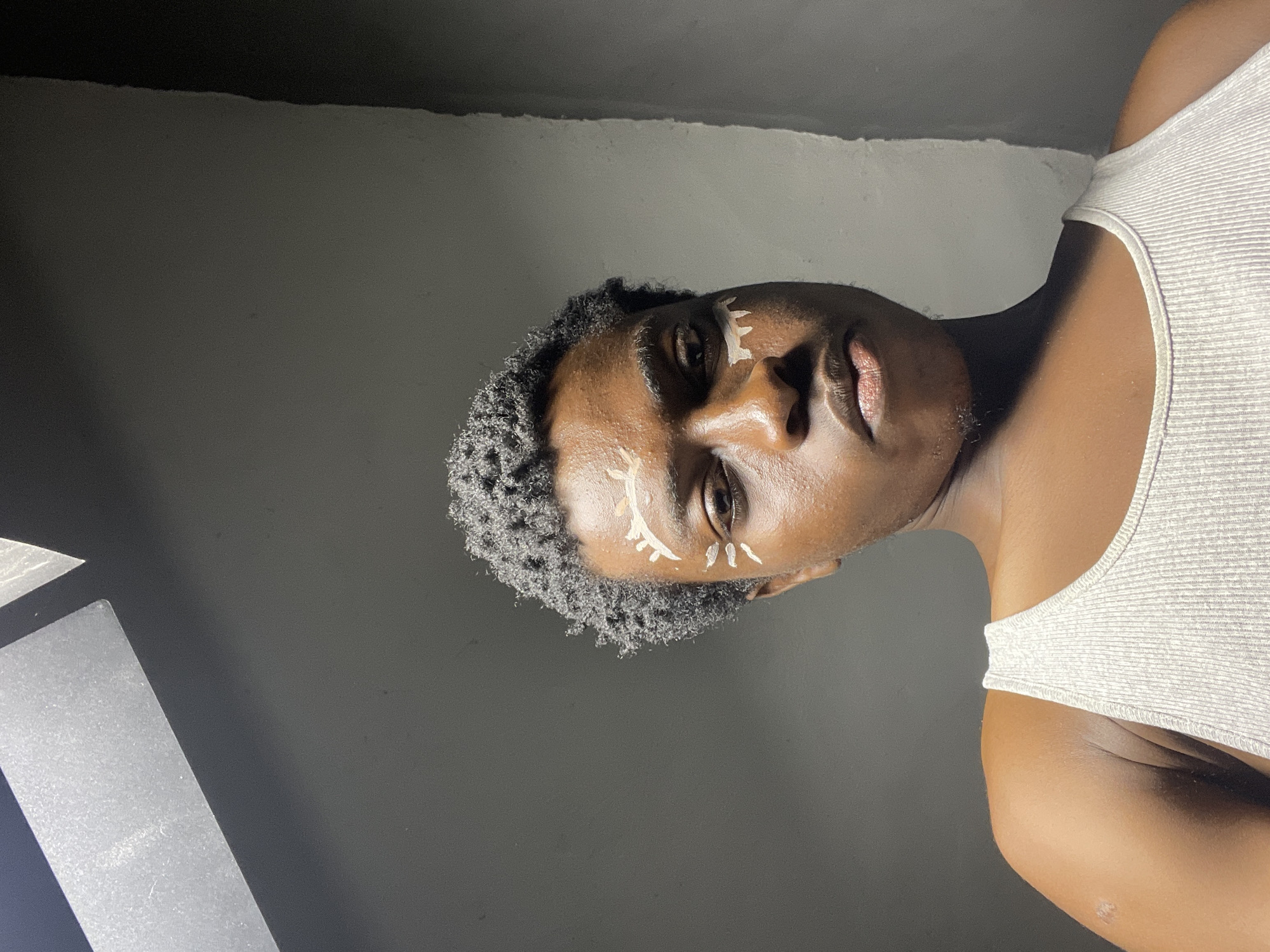A medium shot captures a person with dark skin and short, textured hair from a low angle. They are wearing a ribbed white tank top. White makeup is applied to their face in a stylized pattern around their eyes, resembling sunbursts and eyelids. The background is a dimly lit, gray wall with a rough texture on the right side. Above, a dark, angled structure with a textured surface suggests an overhead element, possibly part of a ceiling or awning. The lighting is dramatic, with shadows emphasizing the contours of the face. The scene appears to be indoors, possibly in Nkpor, Nigeria, given the context of the request.