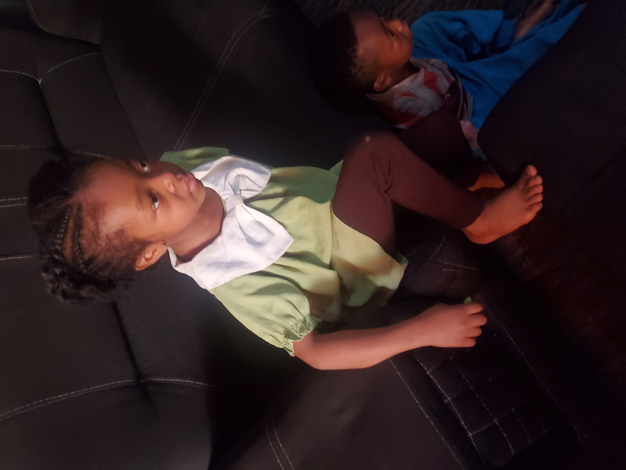 This image, taken indoors in Jos, Nigeria, features two young children resting on a dark-colored sofa. The lighting is dim, casting a soft glow on the subjects.
In the foreground, a child with dark skin and braided hair is lying on their back, looking upwards and slightly to the right. They are dressed in a light green long-sleeved top with a prominent white collar, paired with dark brown or maroon leggings. Their bare feet are visible towards the lower right of the frame. One hand is lightly gripping a small, light-colored object.
Further back on the sofa, partially obscured, is another child. This child is lying on their side, facing away from the camera, and is wearing a bright blue garment over what appears to be a patterned shirt. Their face is not clearly visible.
The sofa itself is dark, likely black or a very dark brown, and features visible stitching details on its upholstery. Both children appear relaxed and comfortable in their positions. The overall atmosphere suggests a quiet moment of rest or casual activity. There is no visible text within the image.
