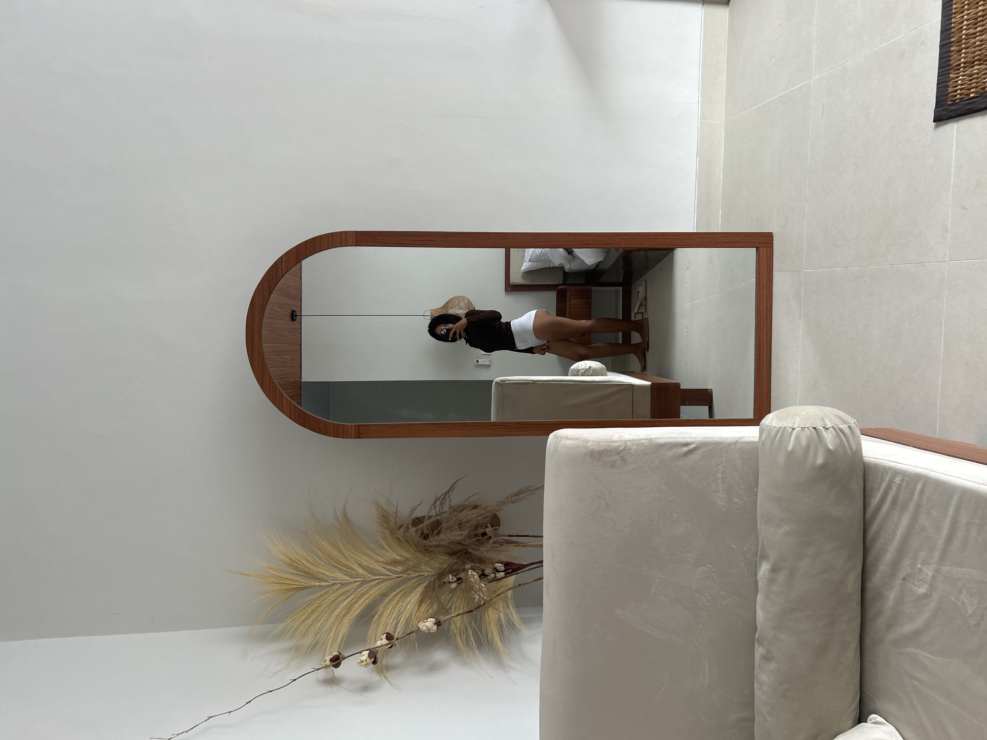 In Abiansemal, Indonesia, a person is captured in a reflection within a tall, arched wooden-framed mirror. They are standing on a light-colored tiled floor, wearing a dark long-sleeved shirt and white shorts, with their back to the viewer. The room features a large, light-colored sofa with a bolster pillow in the foreground. Behind the sofa, a dried floral arrangement is placed against a plain white wall. To the right of the mirror, a bed with white bedding is partially visible, along with a bedside table. The overall environment suggests a well-lit, minimalist interior.