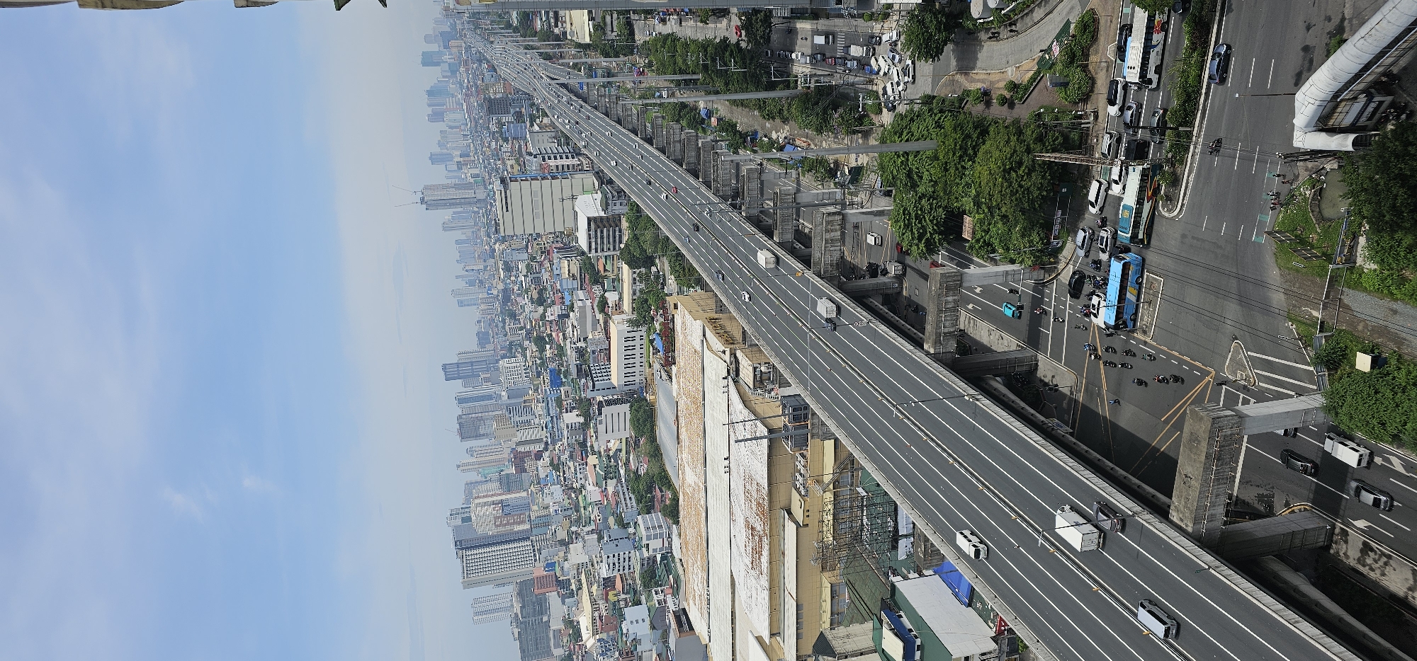 This is an aerial photograph of a highway in Makati, Philippines, under a bright, partially cloudy sky. The highway, elevated on concrete pillars, dominates the foreground and middle ground, with multiple lanes of traffic visible. Vehicles, including cars, trucks, vans, and motorcycles, are present on the highway and on the streets below.
In the background, a dense urban landscape unfolds with numerous high-rise buildings and other structures. Some buildings appear to be under construction, with cranes visible. The streets below the highway are lined with trees and smaller buildings. In the lower right of the image, there is a pedestrian overpass. The overall scene depicts a bustling urban environment with significant infrastructure development.