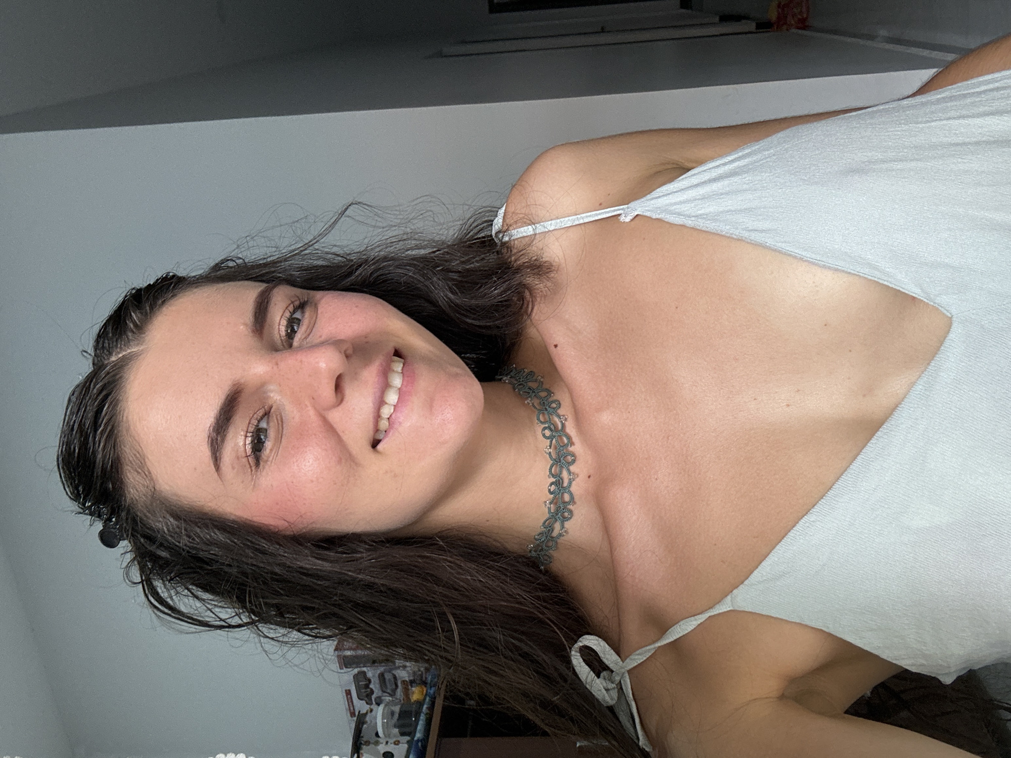 A woman with brown hair smiles at the camera. She wears a light-colored tank top and a lace choker necklace. Part of her dark hair is held back with a black clip. The background is out of focus but suggests an interior space. This image doesn't provide enough information to determine a specific location in Mũi Né, Vietnam.