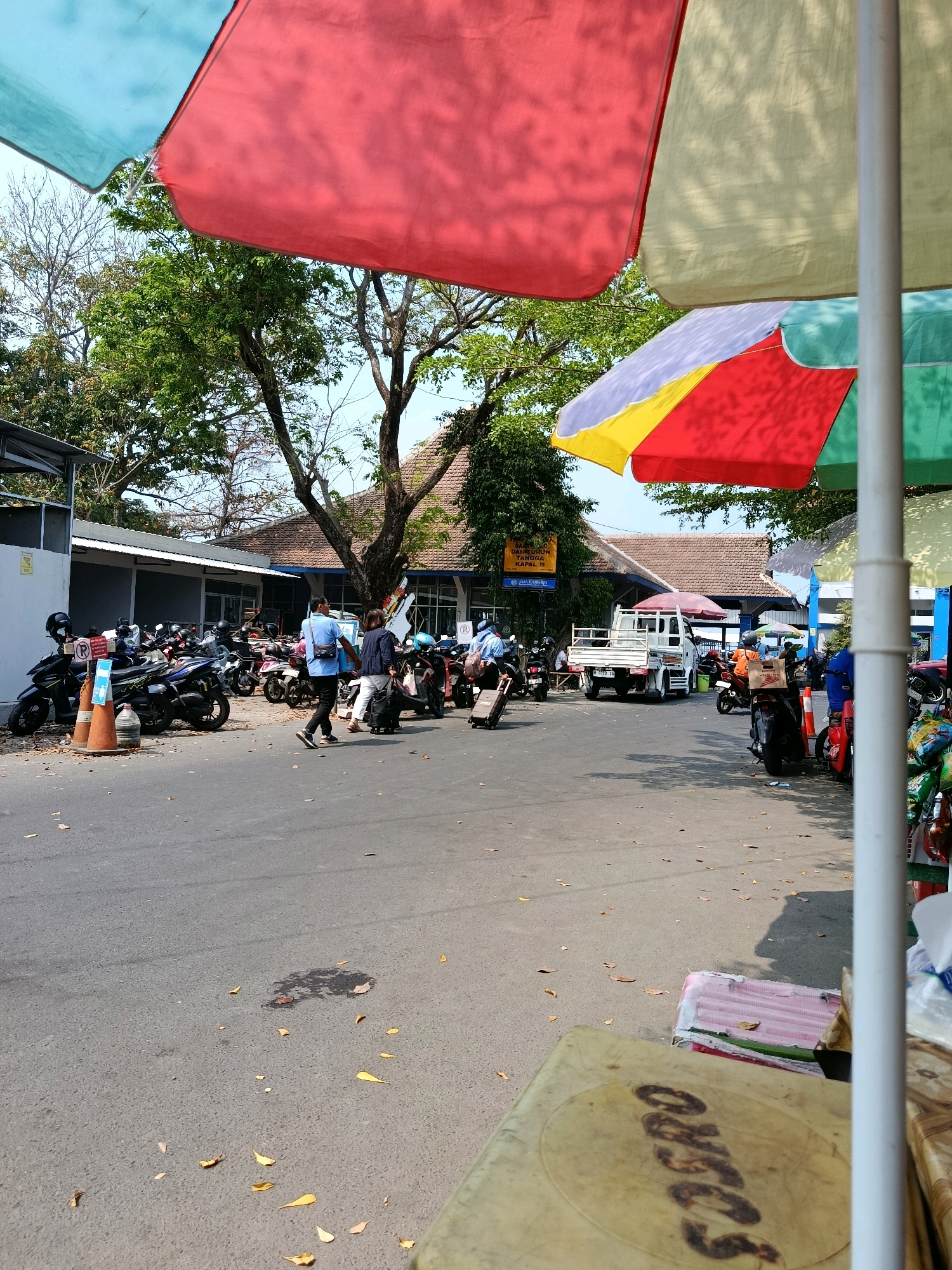 The image depicts an outdoor scene, likely a transportation hub or waiting area, with a visible signage indicating "JASA RAHARJA" which is an Indonesian government-owned insurance company, suggesting a connection to public transport or travel. In the foreground, a weathered, circular table with faded writing is visible. The midground shows several parked motorcycles and a small white truck. People are seen walking, some with luggage, navigating the paved area. Overhead, colorful umbrellas (red, yellow, green, and blue) provide shade. The environment is characterized by scattered dry leaves on the ground and trees with foliage. A building with a tiled roof is present in the background, and additional signage is visible on it. The scene appears to be in Jepara, Indonesia, given the presence of Indonesian signage and the typical architecture and vehicles seen in the country.