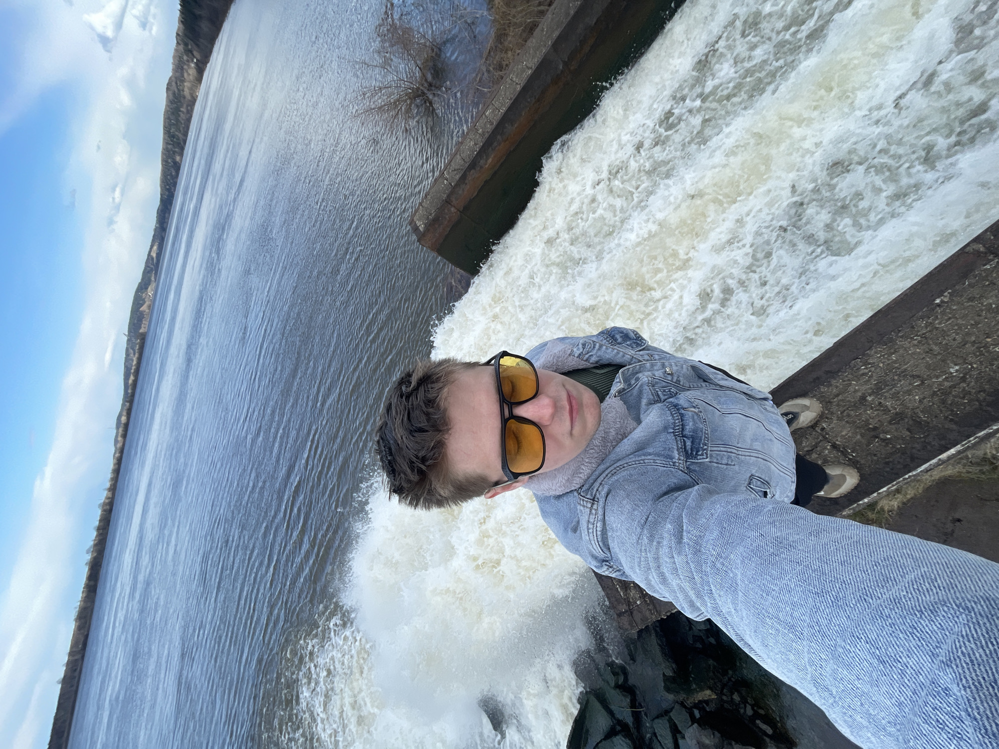 A young person is captured in a selfie taken from a top-down, wide-angle perspective, standing at the edge of a concrete or stone structure overlooking a significant drop where water cascades. The individual, who appears to be a young adult male, is wearing a light blue denim jacket over a green shirt, and light-colored jeans. They have short, dark hair and are wearing distinctive yellow-tinted sunglasses. Their expression is neutral, focused on the camera.
The person is positioned with their back mostly to a large body of water, possibly a river or a reservoir, which stretches into the distance under a partly cloudy sky. On the left side of the frame, the calm surface of this water body reflects the sky. A distant dark treeline or hillside is visible along the horizon. To the right of the person, a large volume of water is intensely white and foamy as it spills over the edge of the structure, creating a waterfall or spillway. The structure itself is made of dark, weathered concrete or stone. The person's feet, clad in light-colored sneakers, are visible near the edge of this structure. The scene conveys an outdoor setting with a prominent man-made water control feature. The time of day appears to be daylight, with diffused lighting. No visible text is present in the image.