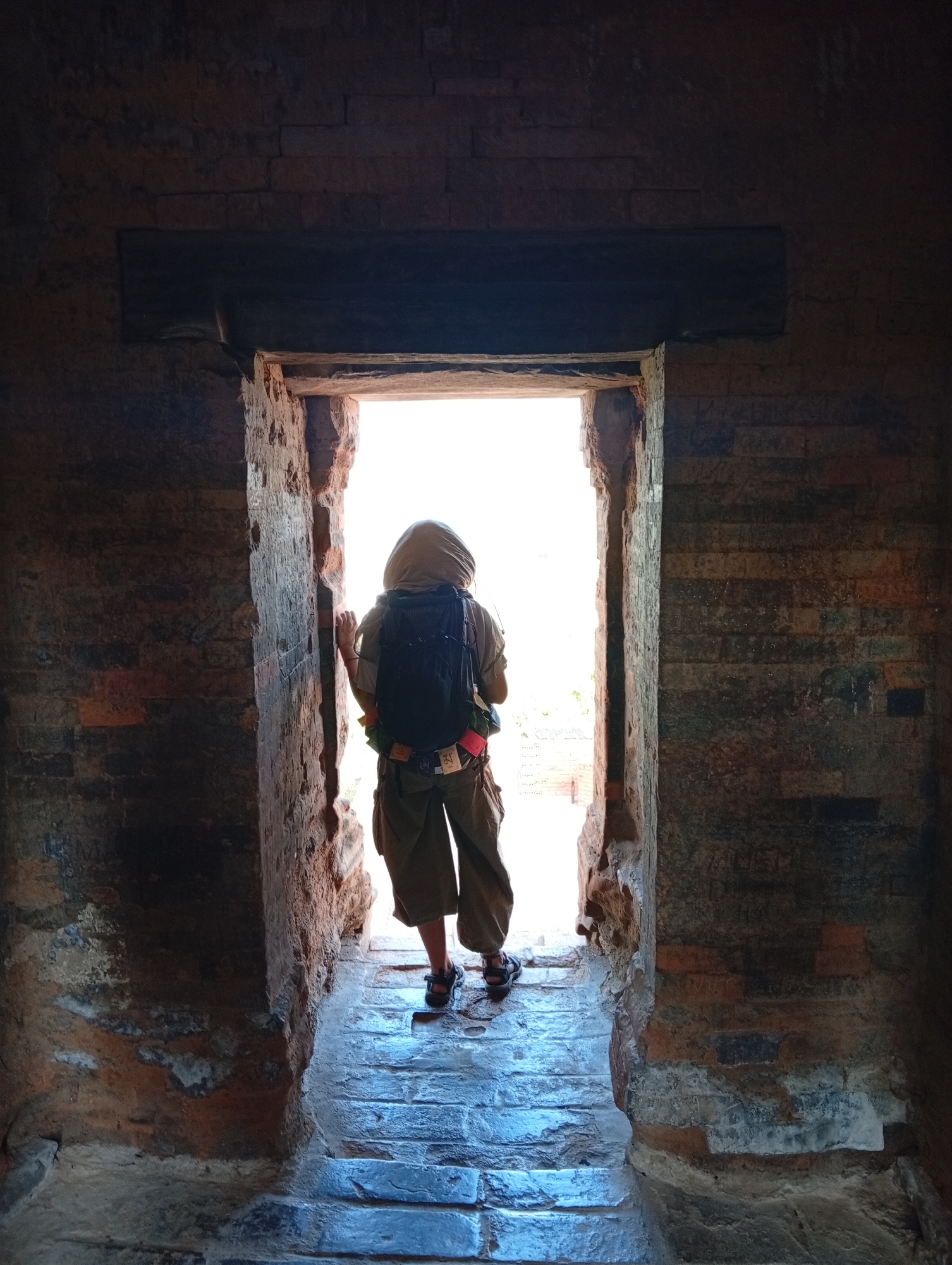 The image depicts a person standing at the threshold of a dark, weathered brick doorway, looking out into a brightly lit exterior. The scene is viewed from within a dimly lit, ancient-looking structure with rough, reddish-brown and dark brown brick walls and a stone floor, possibly an old temple or ruin. A sturdy, dark wooden or stone lintel spans the top of the doorway.
The solitary individual is seen from behind, wearing a light-colored hooded garment that covers their head, khaki or olive-green cropped pants, and sandals. A black backpack is worn, to which several small, colorful items are attached. Specifically, a small red item and a white rectangular tag or piece of fabric with dark, intricate symbols, possibly Tibetan script, are visible on the lower right side of the backpack.
The person stands facing outwards, with their hands lightly touching the doorframe, suggesting a moment of pause or contemplation before stepping into the bright, sunny environment outside. The intense light flooding through the doorway indicates it is daytime, likely a clear and sunny day. The architectural style, characterized by aged brickwork and a substantial threshold, points to a historical site. The context of Phan Rang – Tháp Chàm, Vietnam, suggests this could be one of the region's ancient Champa towers. The mood conveyed is one of quiet exploration and anticipation.