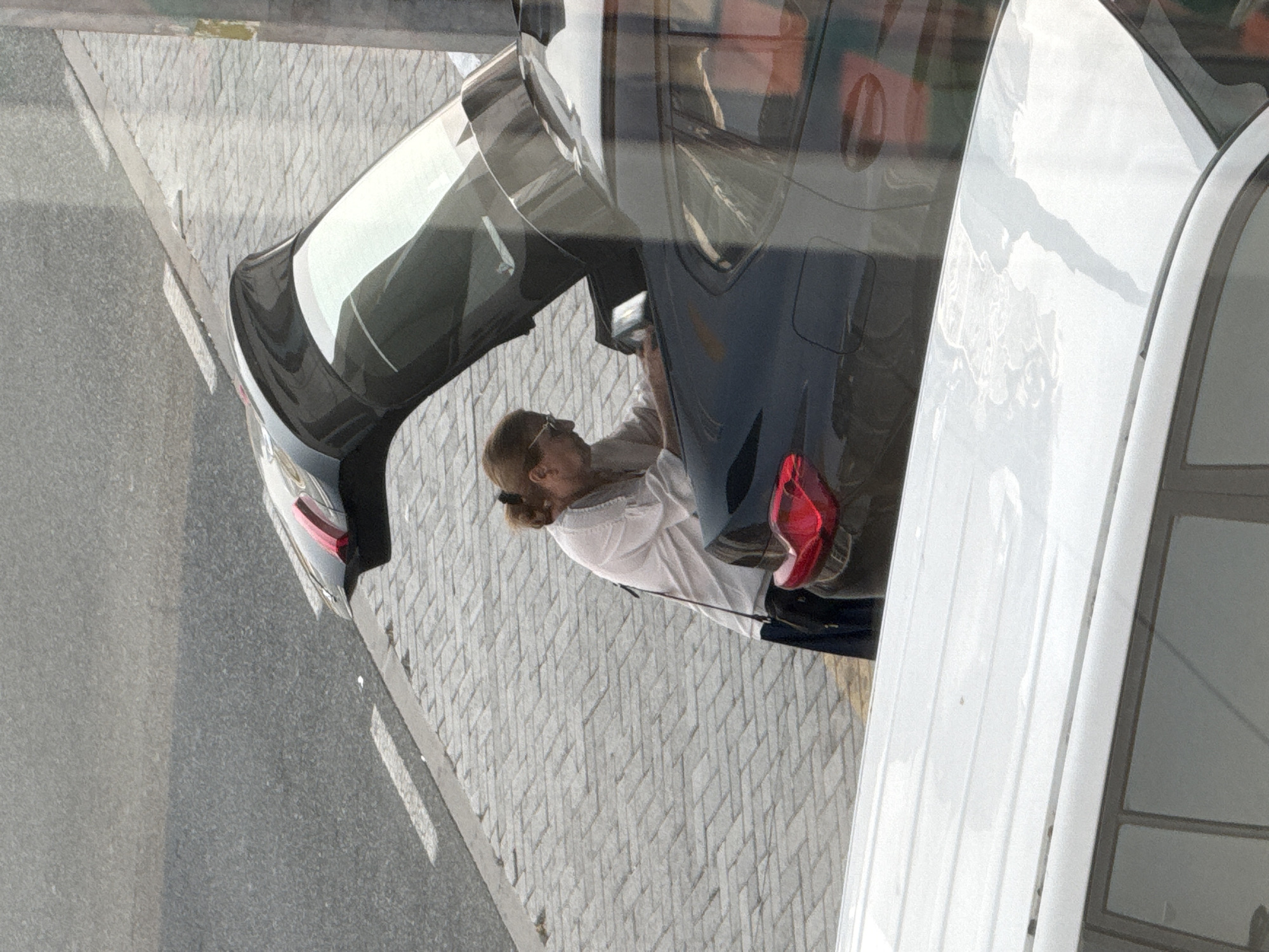 A medium shot captures a woman with her hair in a bun, wearing glasses and a white blouse, bending over the open trunk of a dark-colored car. She appears to be placing or retrieving an item from the trunk. The car is parked on a paved area adjacent to a gray asphalt road with white lane markings. A white vehicle is visible in the foreground, partially obscuring the scene. The background shows a textured paved surface and a light pole. The lighting suggests it is daytime. The scene appears to be a street or parking area in a city, consistent with urban environments in Dubai, United Arab Emirates.