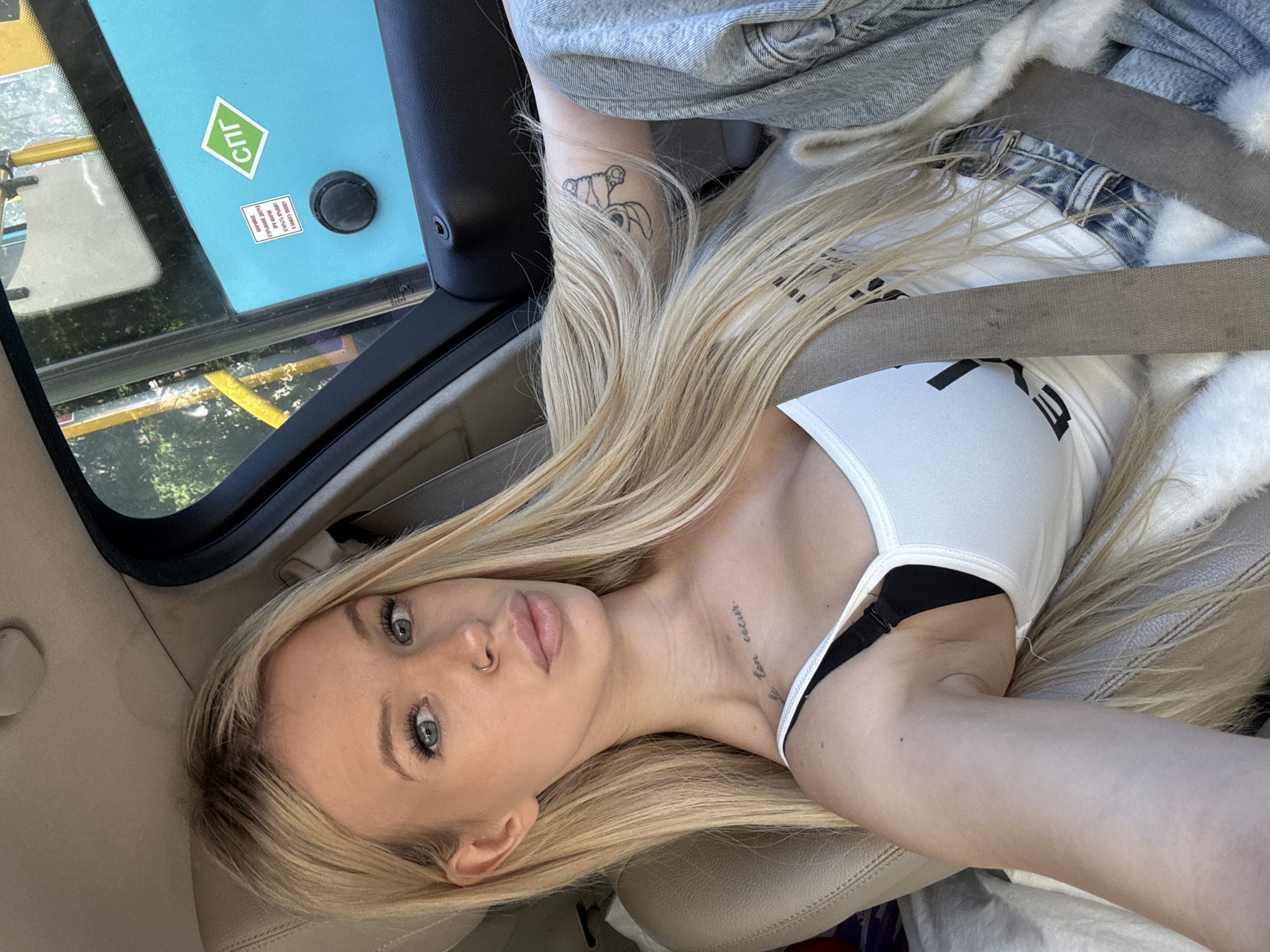 This is a selfie taken by a young woman with long blonde hair. She is sitting in the passenger seat of a car. She is wearing a white tank top with a black bra underneath, and a denim jacket lined with white fur. A seatbelt is visible across her chest. She has light blue eyes and is looking at the camera. There are two tattoos visible: one on her left forearm and another small one on her neck.
In the background, through the car window, there are trees and a yellow structure. On the window itself, there are two stickers. One is a green diamond shape with white text that appears to be Cyrillic. Below it is a smaller rectangular sticker with red and white text, also in Cyrillic. A black circular object is also visible on the window.
The lighting suggests it is daytime, with bright sunlight filtering in. The overall mood is casual and relaxed, as if she is on a car journey.
The visible text on the stickers is:
- On the green diamond: "ГПЛ"
- On the smaller rectangular sticker: "ВАЖНО! ПРИ НЕИСПРАВНОСТИ", followed by smaller text that is difficult to read.