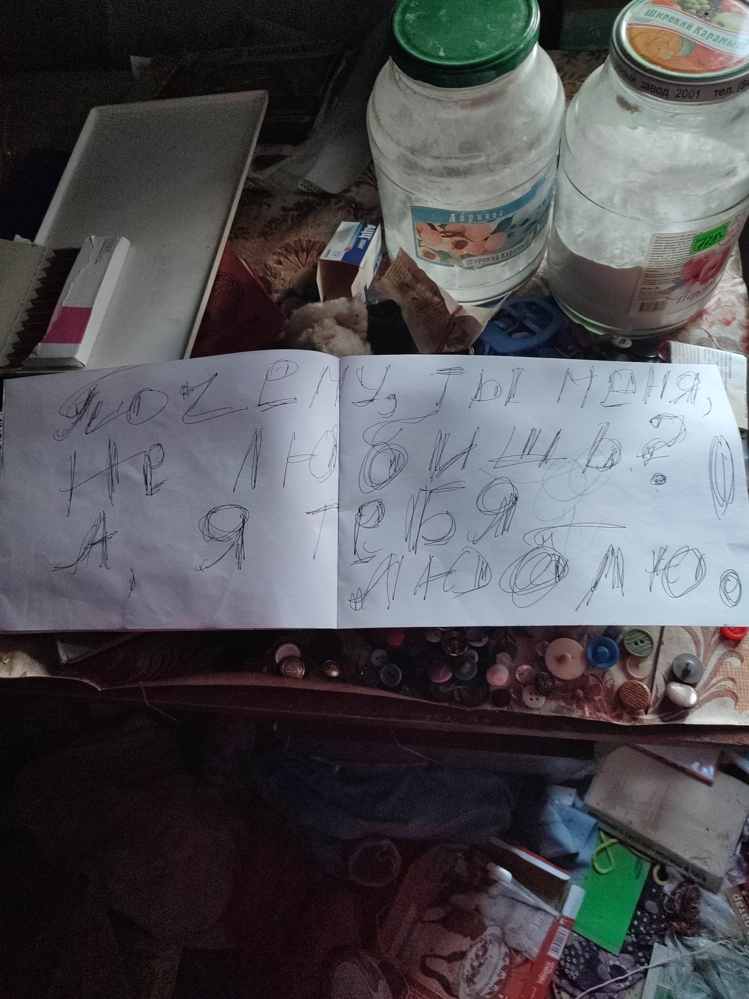 The image shows a cluttered, indoor scene with a handwritten note on white paper as the central focus. The note contains text in Russian, appearing to be a message written with a black pen. The surrounding environment is disorganized, with various objects scattered on a table and in the background.
On the table, to the left of the note, there is a white rectangular object and a partially visible box with pink and white elements. To the right of the note, there are two large glass jars, one with a green lid and the other with a silver lid. Both jars appear to contain a white powdery substance. A label on the jar with the green lid has Russian text and an image of apricots, reading "Абрикос" (Apricot) and "Широкий Карамы" (Wide Karamys). The jar with the silver lid also has a label with Russian text and an image of peaches.
In the background, other items are visible, including what appears to be a patterned tablecloth or fabric, and some blurred objects that could be containers or other household items. Below the table, the image shows a dark area filled with various items, including buttons of different sizes and colors, fabric scraps, and other debris.
The image does not contain any people or visible actions or interactions. The environment suggests a storage area or a less-used part of a home. Without further information or context, it is not possible to determine the exact location within the city of Gremyachinsk, Russia, other than inferring it is an indoor setting within that city.