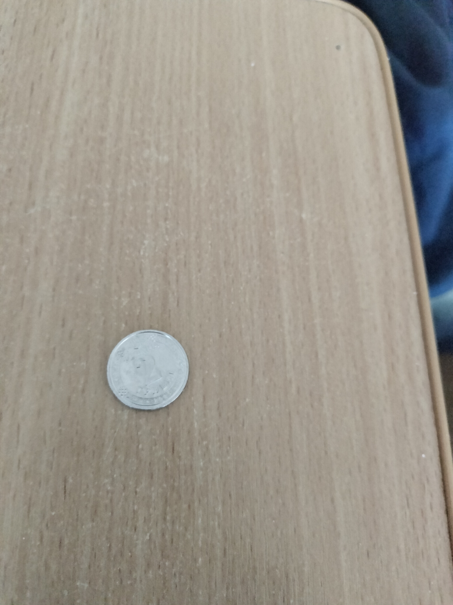 A single silver-colored coin rests on a light brown, wood-grained surface. The coin appears to be circular with raised markings or a design on its surface. The background surface exhibits a subtle wood grain pattern and some light-colored speckling, possibly dust or debris. To the right of the surface, a portion of dark blue fabric is visible, suggesting the surface might be a table or desk with clothing nearby. The image does not provide any information about the city of Chernihiv, Ukraine, or any specific location within it.