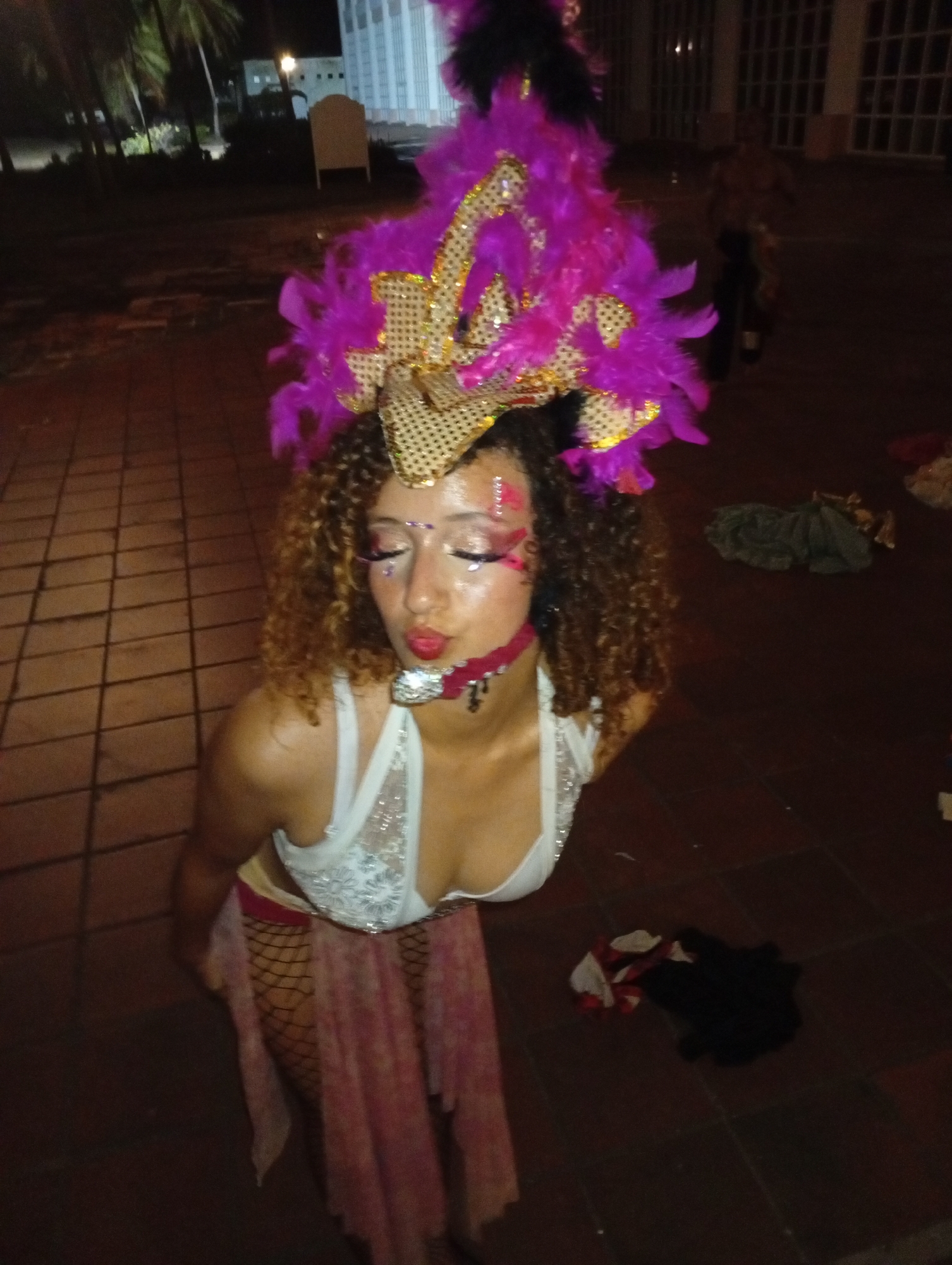 A woman in a revealing outfit and elaborate headdress is kneeling on a tiled surface. She wears a white top with lace detailing, a pink skirt over fishnet stockings, and a large headdress featuring gold accents and bright pink feathers. Her makeup includes glitter and designs around her eyes. She also wears a choker. In the background, there are palm trees, a building with illuminated windows, and other indistinct figures. The scene appears to be outdoors at night. The location may be Pedro González, Venezuela.