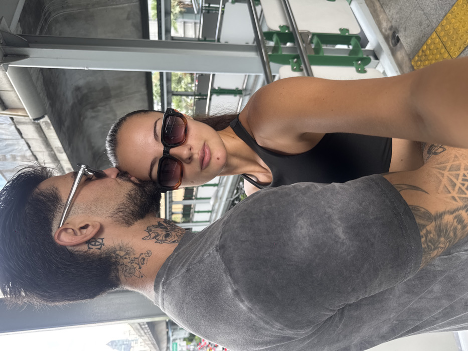 The image displays two individuals on an elevated platform, identified as a Bangkok BTS Skytrain station based on the distinct elevated concrete structure, metal railings with green and white panels, and yellow tactile paving. In the foreground on the left, a male subject is partially visible from the side, wearing a faded grey t-shirt and silver-framed sunglasses. He has visible tattoos on his neck, including geometric patterns and a stylized mask-like figure, and additional tattoos on his arm. He is leaning down, performing an action consistent with kissing the forehead of the female subject. The female subject is positioned on the right, facing the camera directly, suggesting she is operating the device for a selfie. She wears a black athletic top and large, square-framed sunglasses with brown gradient lenses. Her dark hair is pulled back. Her left arm is extended towards the camera. The background features distant urban buildings and a street level with visible traffic, consistent with a city environment like Bangkok.