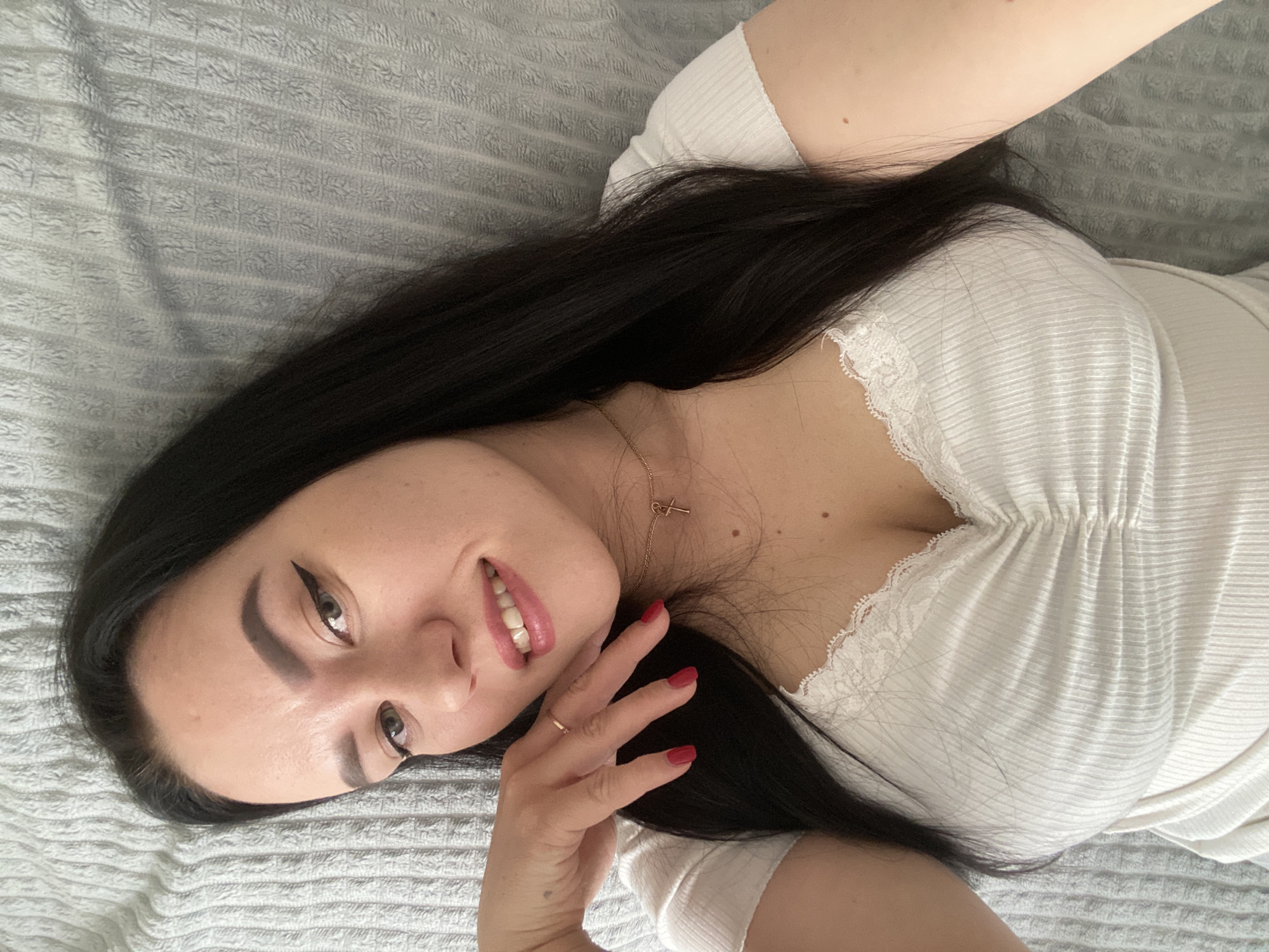 A young woman is depicted lying down on what appears to be a bed or couch, covered by a light grey, textured blanket or bedspread. She is positioned diagonally across the frame, looking towards the camera with a relaxed smile, her lips slightly parted. Her long, dark hair is spread around her head and shoulders.
She is wearing a light-colored, possibly white or off-white, ribbed top featuring lace trim along the V-neckline. A delicate gold chain necklace with a small cross pendant rests on her chest. Her fingernails are painted red, and she wears a thin gold ring on a finger of her right hand, which is gently placed near her face. Her facial features include dark, well-defined eyebrows and visible winged eyeliner. The lighting is soft and even, suggesting an indoor setting during the daytime. The overall impression is one of calm and contentment. No visible text is present in the image. The location context is Chaikovsky, Russia.