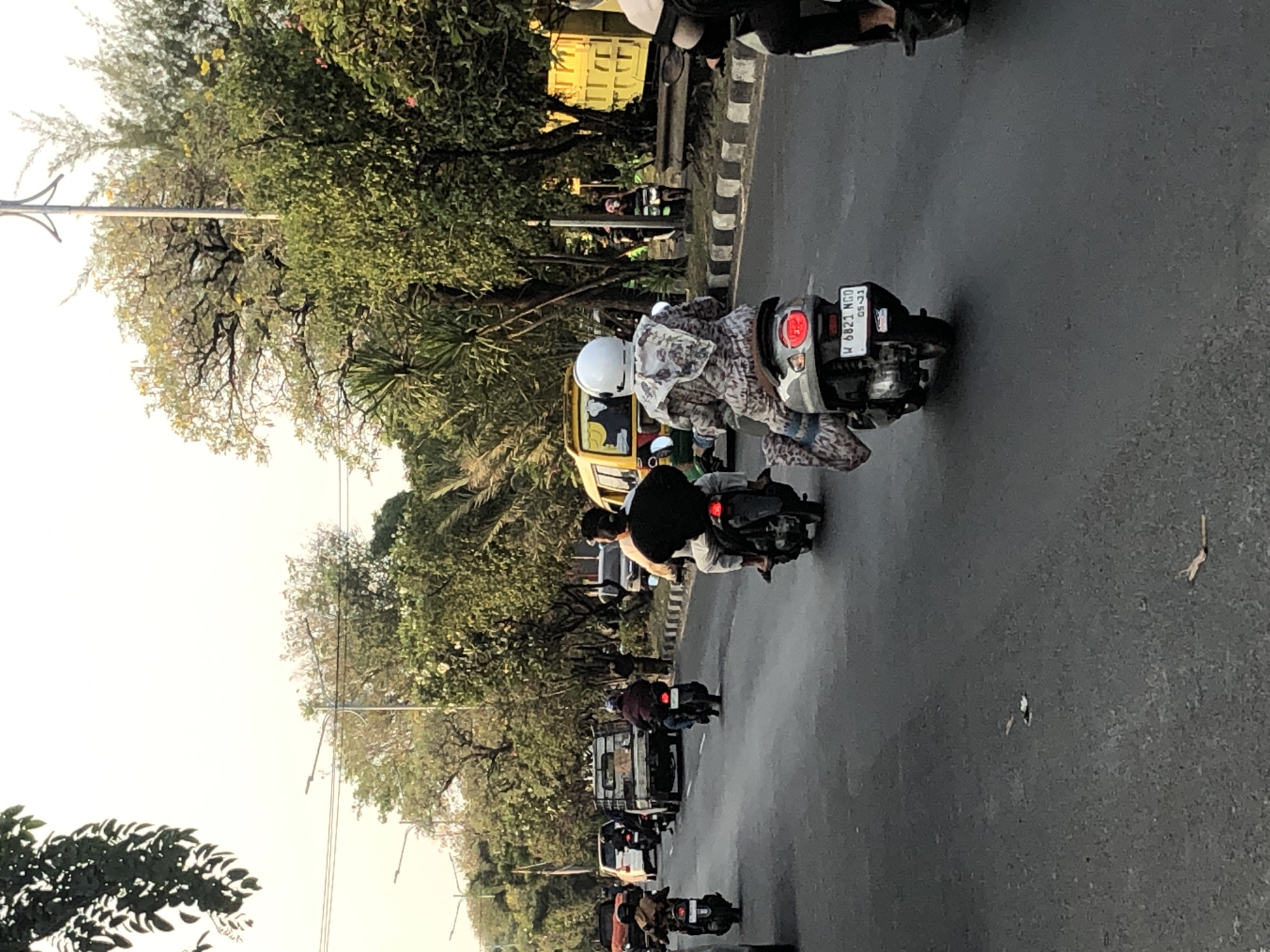 The image depicts a street scene in the city of Ngaban, Indonesia. In the foreground, two individuals are riding motorcycles on a paved road. The motorcycle on the right has a license plate that reads "W 6821 NGO 05-23". On the left, another motorcycle rider is visible, partially obscured. In the middle ground, several other vehicles are present, including a small truck, a yellow van, and cars. The road is lined with trees and foliage, suggesting an urban or suburban environment. The sky appears overcast or hazy.