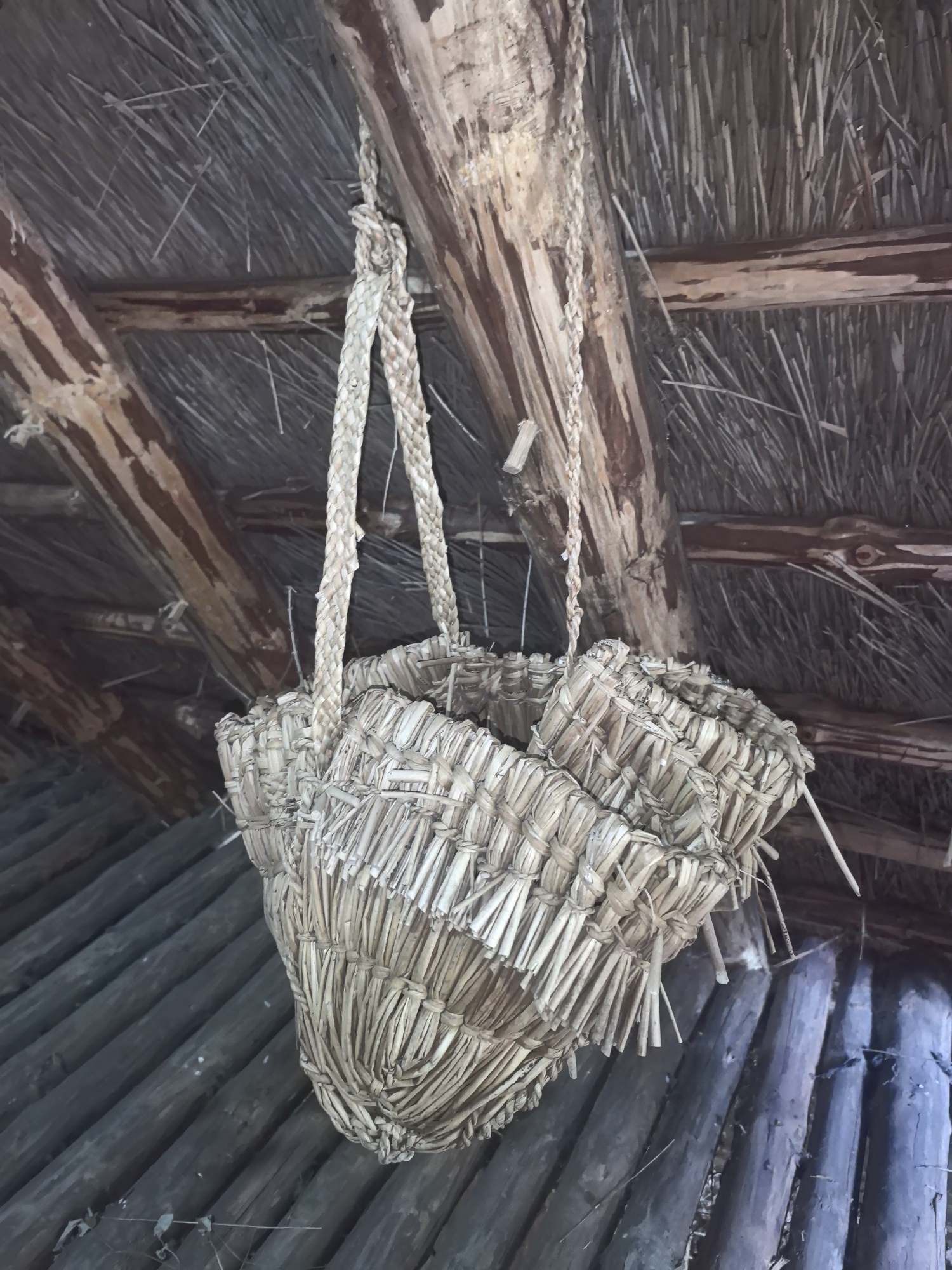 An upward angled medium shot captures a woven straw basket hanging from a rustic wooden ceiling. The basket, composed of dried stalks tied together, has a wide, irregular opening and a braided handle. The ceiling is constructed of rough-hewn wooden beams and a thatched roof made of straw. Below the ceiling, a series of dark, roughly cut logs are visible, arranged diagonally. The scene appears to be in an outdoor or open structure, possibly a shed or primitive dwelling, located in the Камянюцкі сельскі Савет, Belarus. The lighting is natural, casting shadows and highlighting the textures of the materials. There are no people visible in the image.