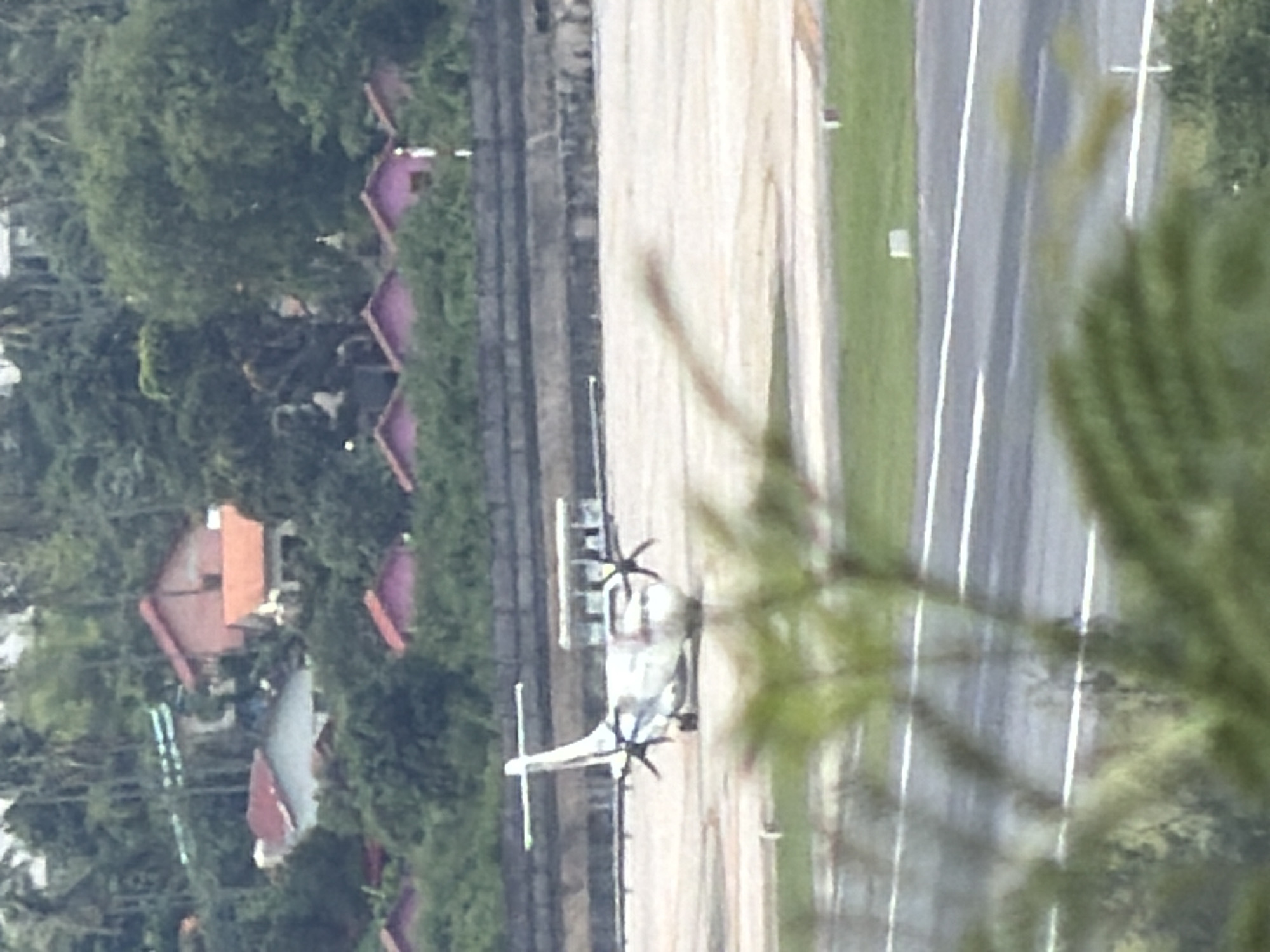 The image captures an aerial view of an airport tarmac in Ban Bang Rak, Thailand, with a turboprop airplane positioned on the runway. The aircraft, primarily white with a visible tail fin and two propellers, is facing away from the camera. In the background, several buildings with red and pink roofs are nestled amongst lush green trees. A concrete structure, possibly a tiered seating area or embankment, runs along the perimeter of the tarmac. The foreground is partially obscured by blurred green foliage, suggesting the photo was taken from a vantage point overlooking the airport.