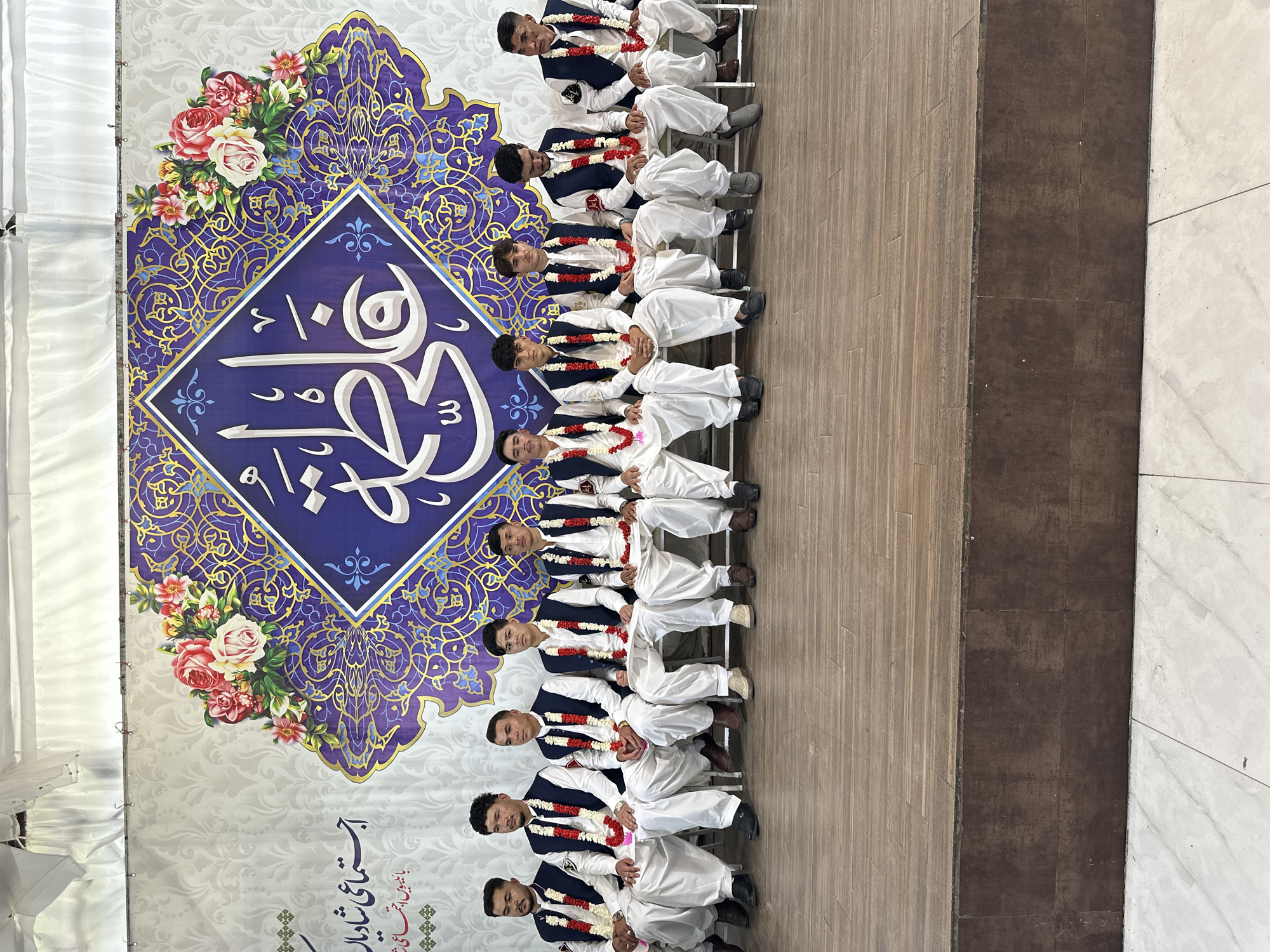 A photograph shows a row of nine people sitting in front of a large backdrop. The individuals are all dressed in matching outfits that consist of white pants and a dark vest, along with a flower garland around their necks. They appear to be posing for a group photo.
The backdrop behind them is a patterned design with a large blue square containing an Arabic or Urdu calligraphic design. Decorative floral arrangements are placed on either side of the square.
The photo appears to have been taken indoors on what looks like a tiled floor. The location is labeled as Unknown, Unknown, and no specific details about the city can be inferred from the image. The setting seems to be related to some kind of event or ceremony.