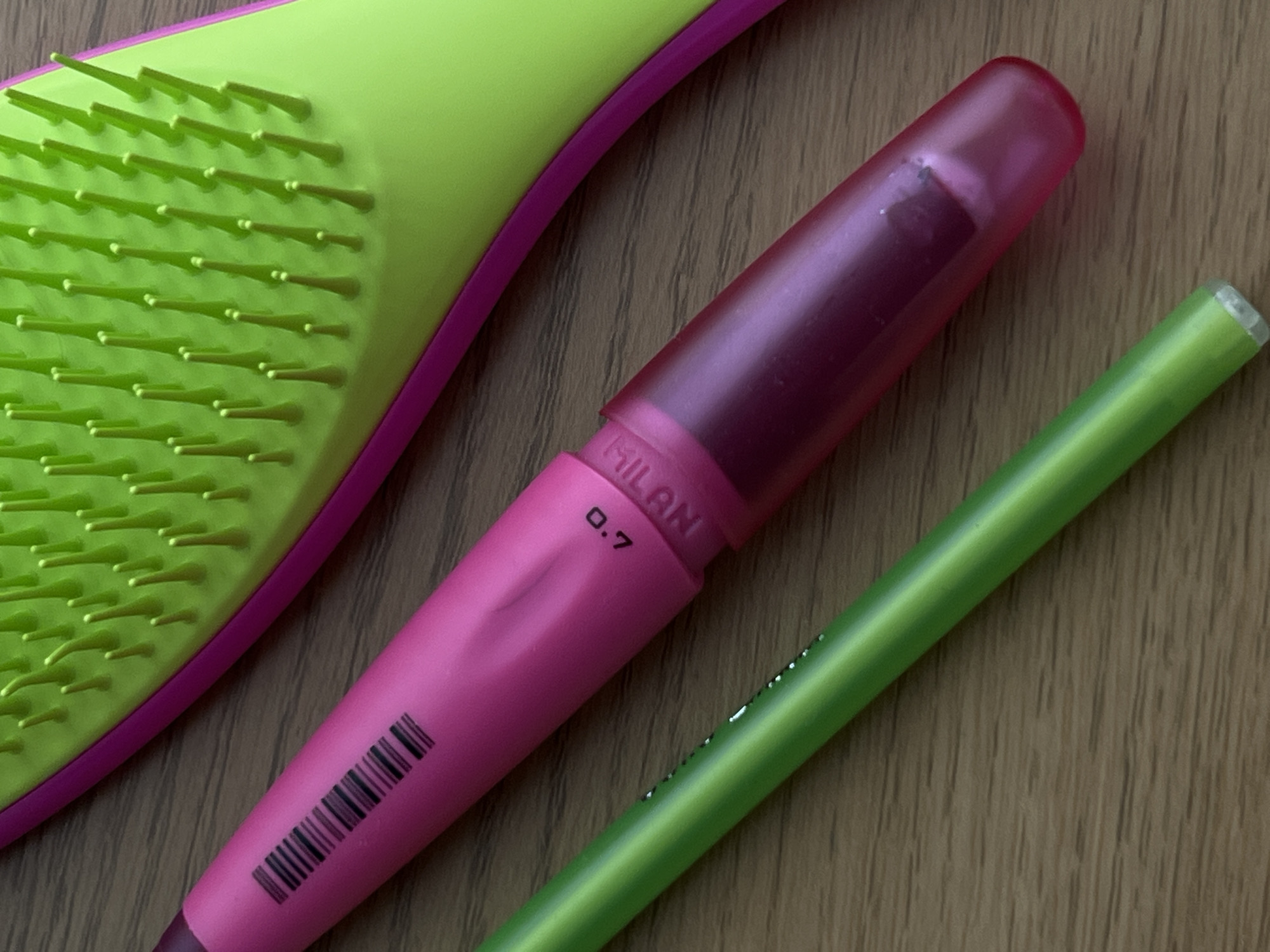 A flat lay photograph captures a collection of colorful everyday items resting on a light brown wooden surface. The arrangement includes a hairbrush, a mechanical pencil, and a pen or pencil.
In the upper left quadrant, part of a hairbrush is visible, featuring numerous bright green, flexible bristles set against a vibrant magenta-pink base. The bristles are uniformly spaced and appear slightly rounded at their tips.
The central subject is a pink mechanical pencil. It has an opaque pink barrel with a transparent, darker pink cap that covers the eraser mechanism. On the barrel, the word "MILAN" is faintly visible, along with the numerical inscription "0.7", likely indicating the lead thickness. A barcode pattern is also printed near the lower end of the pencil. There are some minor indentations or wear marks on the pencil's surface.
Parallel to the pink mechanical pencil and extending along the lower right side of the frame is a slender, bright green writing instrument, which could be another pencil or a pen. Its tip appears slightly worn or discolored.
The background consists of a wooden texture, characterized by subtle grain patterns in varying shades of light brown and beige. The lighting is even, suggesting an indoor setting without direct harsh sunlight. No activity is depicted; the items are stationary. While the image is provided with the location context of Budva, Montenegro, no specific visual elements within the photograph itself confirm this geographical detail.