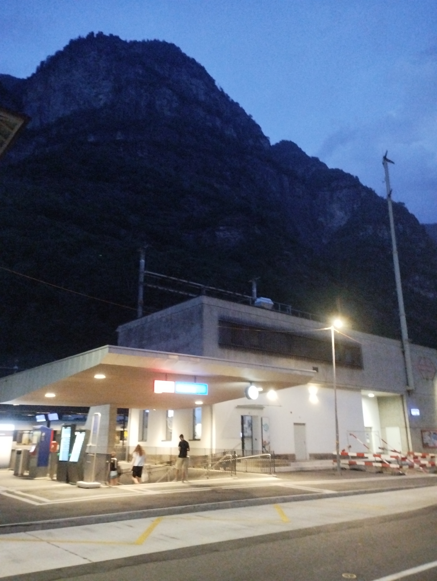This is a full, slightly low-angle shot of a train station in the evening, set against a dark mountain range. The sky is a deep blue with hints of twilight. The station itself has a modern, open-air design with a light-colored canopy extending over a paved area. Visible on the left are ticket machines or information kiosks with illuminated screens. Two people, a woman in a white top and black skirt and a man in a black shirt and light shorts, are standing near stairs leading into the station building. The building is light-colored and features large windows and a prominent lamppost with a bright light. To the right of the building, there are barriers with red and white diagonal stripes, suggesting ongoing construction or a restricted area. A tall utility pole with a crossbar stands to the right, reaching towards the sky. The overall impression is that of a functional, contemporary transportation hub at dusk, framed by the dramatic natural backdrop of the mountains.