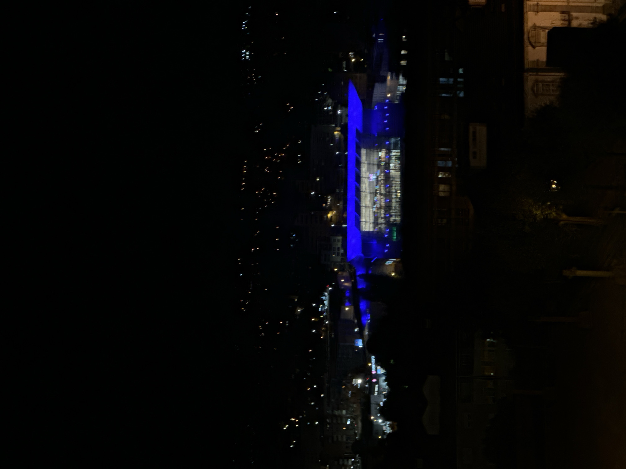 The image captures a nighttime view of Vila Real, Portugal, from a slightly elevated perspective. The scene is dominated by a prominent, modern building on the right side, which features a striking, brightly illuminated blue facade. This building appears to be made of glass and steel, with numerous windows that show interior lights, contributing to its luminous presence against the dark sky.
To the left of the blue building, the city stretches into the distance, marked by a scattering of small, twinkling lights. These lights, varying in color from white to warm orange, suggest streetlights, vehicle head/tail lights, and lights from other buildings, illustrating the urban landscape at night.
Further to the right and slightly behind the blue structure, the outlines of other buildings are visible, though much darker and less distinct. One appears to be an older, more traditional architectural style, identifiable by its roofline and darker facade. The immediate foreground and far left of the image are largely obscured by darkness, possibly indicating unlit areas or trees.
The time of day is clearly night, with no discernible natural light. The weather appears clear, with no visible precipitation or heavy cloud cover. There is no specific activity or event captured, rather a static panoramic view of the illuminated city. No legible text is visible within the image.