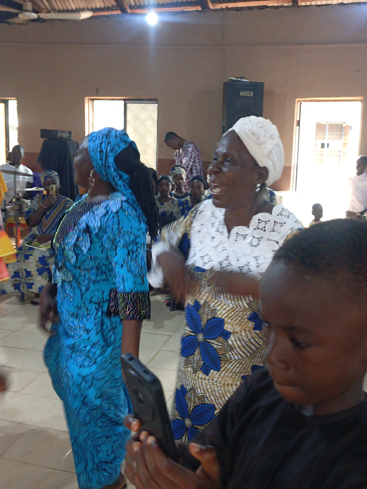 The image shows a gathering of people, possibly a celebration or event, inside a building in Akwanga, Nigeria. The interior features tiled floors, light brown walls, and visible windows and doorways letting in natural light. A ceiling fixture provides additional illumination.
Several women are prominently displayed, wearing vibrant, patterned dresses and head coverings. One woman in the foreground wears a blue dress and headscarf, while another woman wears a white head covering with a patterned dress featuring blue floral designs. Other women, similarly dressed, are seated in the background, some holding phones, possibly recording the event. A group of seated children are visible as well.
A young boy is in the foreground, holding a phone and looking down at the screen. Behind the group, a man stands wearing a patterned shirt, and other individuals stand near the doorway in the background. A dark speaker box is visible against the wall.