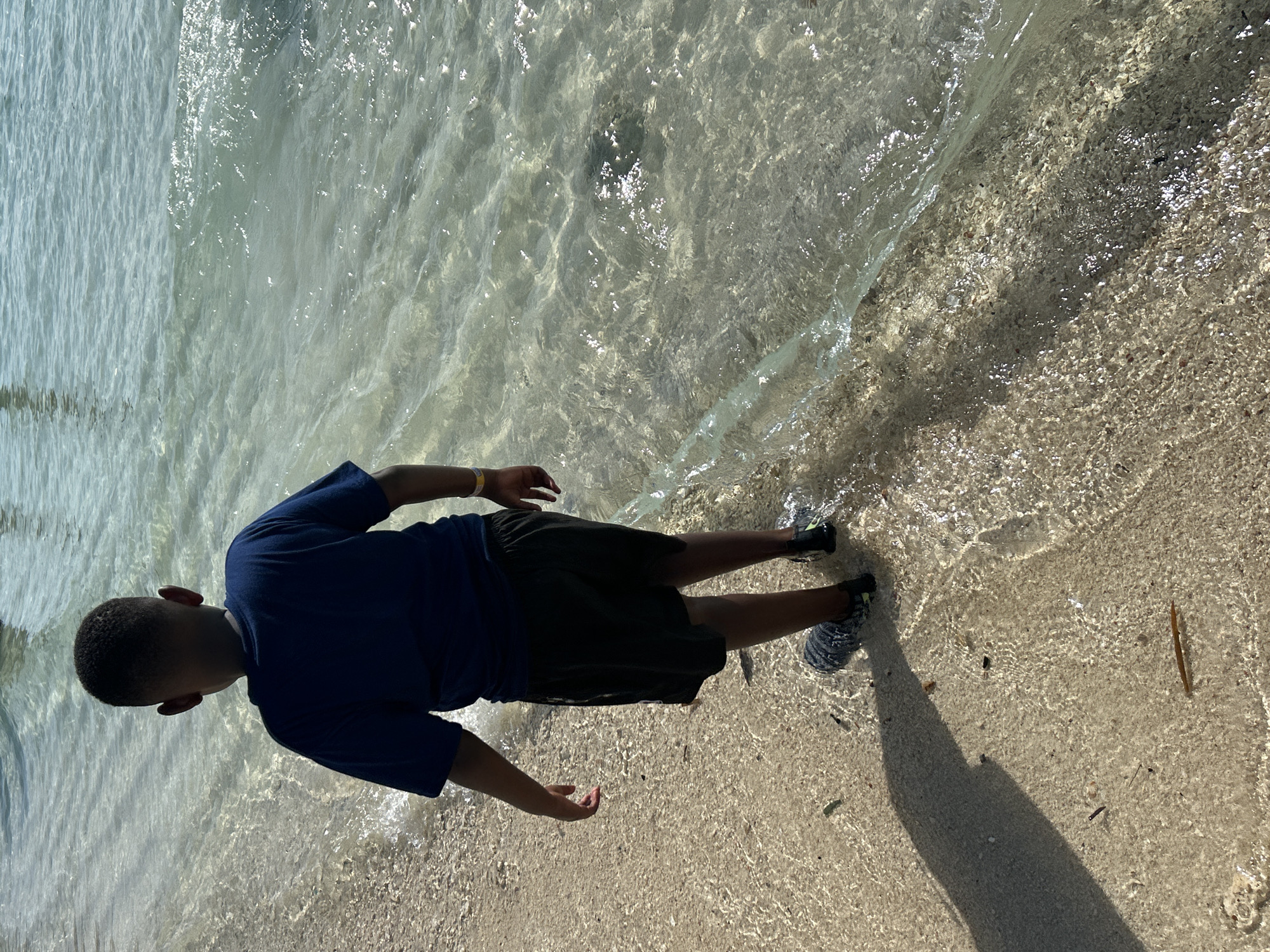 An overhead, medium shot shows a young Black boy from behind, wading in the shallow, clear water of a beach in Phru Nai, Thailand. He is wearing a dark blue t-shirt, dark shorts, and water shoes. The boy's right arm is bent and his hand is near his hip, while his left arm hangs loosely by his side. His shadow stretches across the sand to his left. The water is calm and shallow, with small waves gently lapping at the shore. The sand is light brown and speckled with small pebbles and shells. The sun shines brightly, creating reflections on the water's surface.