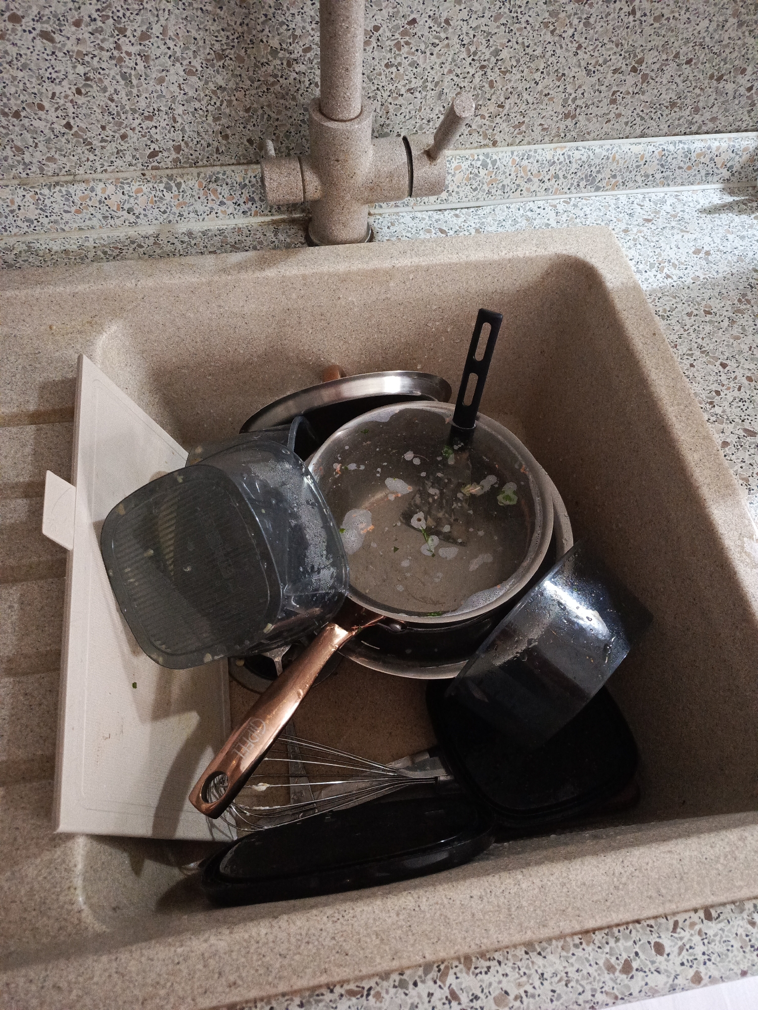 The image shows a beige kitchen sink filled with dirty dishes. The sink is located under a beige faucet set against a wall with a speckled beige and brown pattern. Inside the sink are various items, including a white cutting board, a black air fryer basket, a pot with a black handle and lid, a frying pan, a whisk with a copper handle, and several black lids. Food residue is visible in the pot. The location is not identifiable in the city of Северный, Russia.